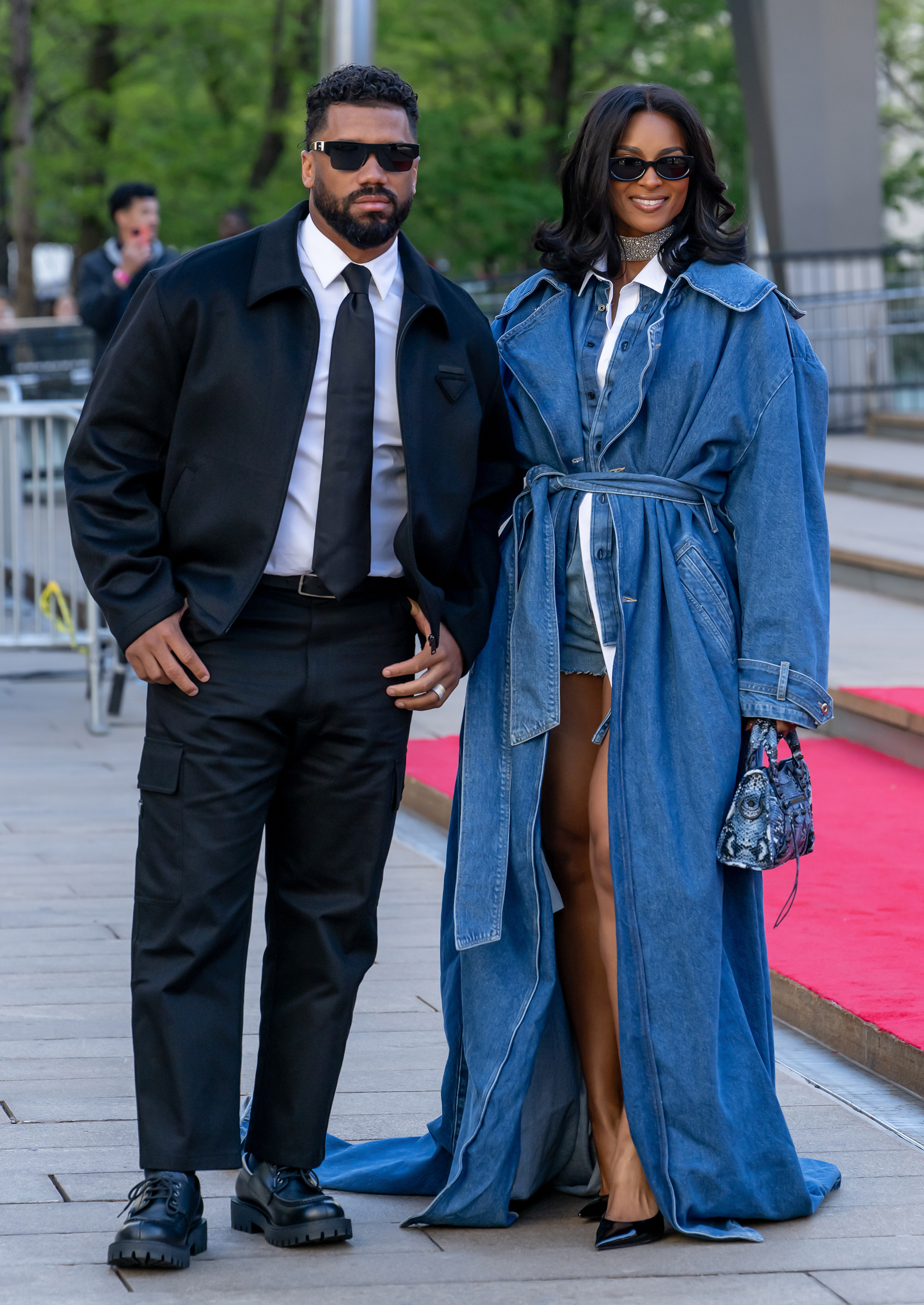 Russell Wilson and Ciara stand closely side by side along the sidewalk, both wearing dark sunglasses that add a cool, composed edge. Her belted denim coat drapes elegantly as she carries a patterned bag, while his structured black outfit keeps the look sharp and understated.
