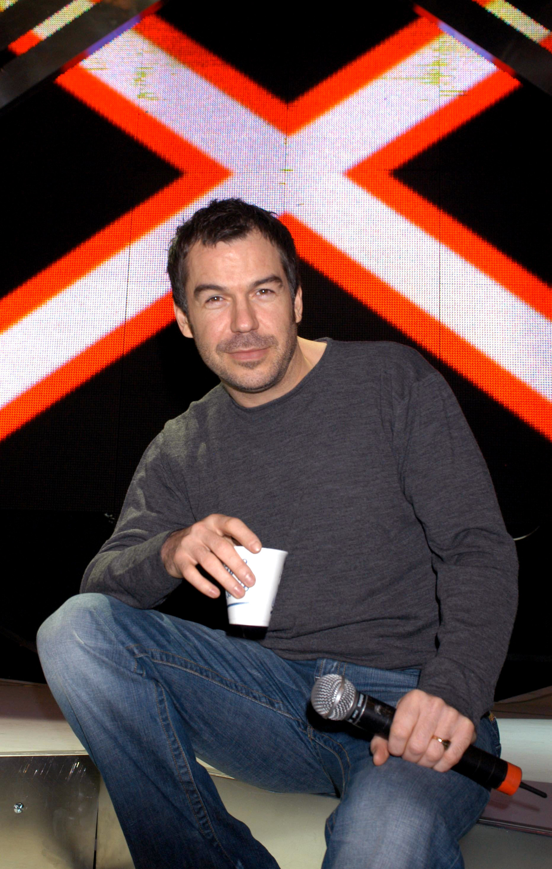 Brookstein perches casually on a ledge during rehearsals at the London Arena in February 2005, jeans-clad and holding both a microphone and a paper cup, with the illuminated "X Factor" logo blazing behind him.