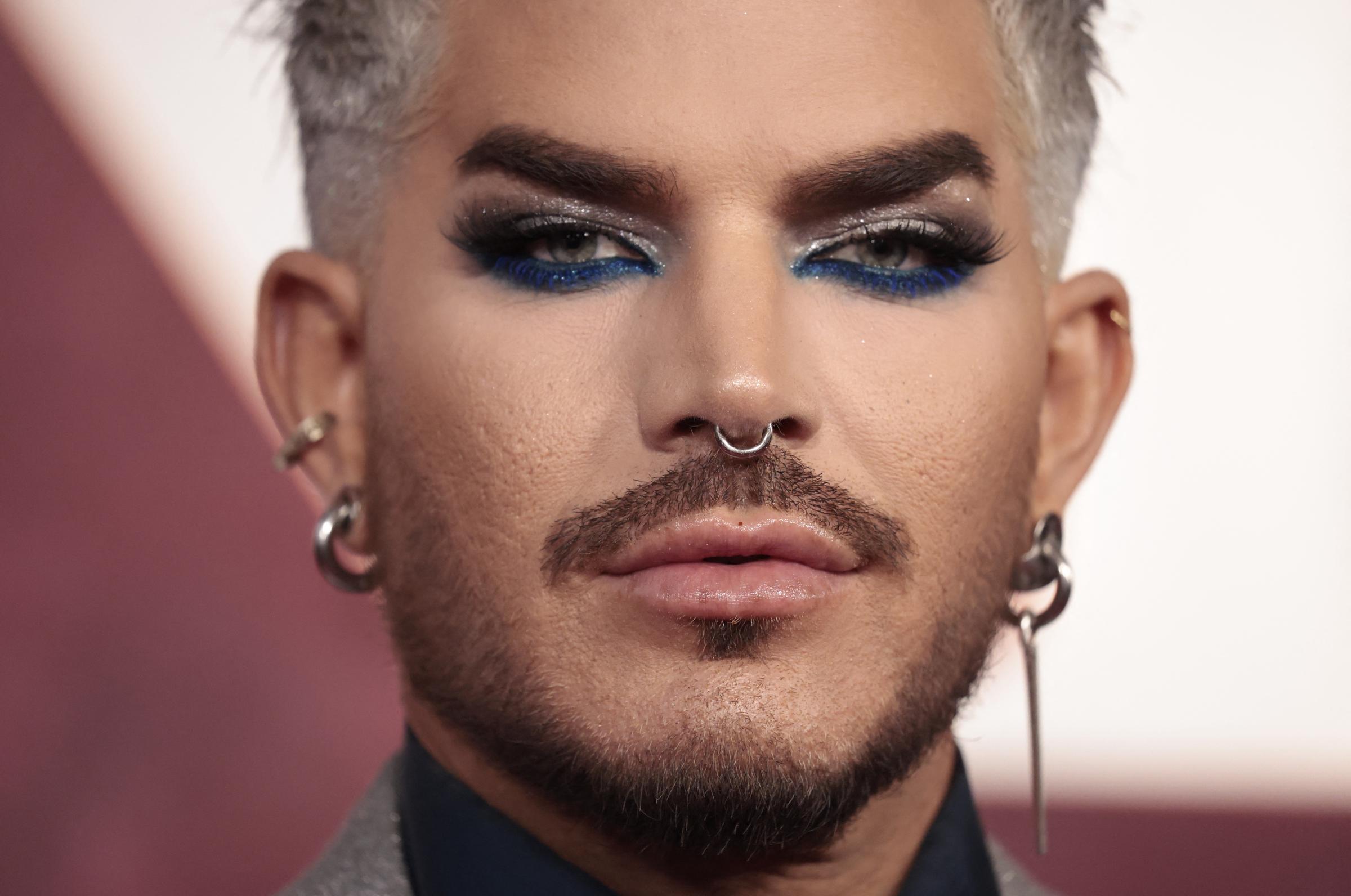 A close-up captures Adam Lambert's piercing gaze framed by electric-blue liner, glitter-shadowed lids, and voluminous lashes. His silver hair, septum ring, and detailed facial grooming amplify a daring look rooted in glam, drama, and confidence.