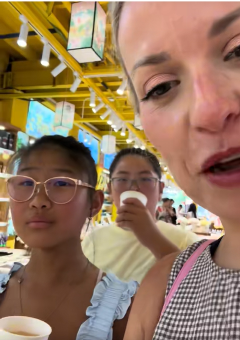 Amanda "Mandy" Karolkiewicz stands with her two adopted daughters as they hold small sample cups inside a brightly lit market or food hall during their trip to China. | Source: TikTok/mandykarolk
