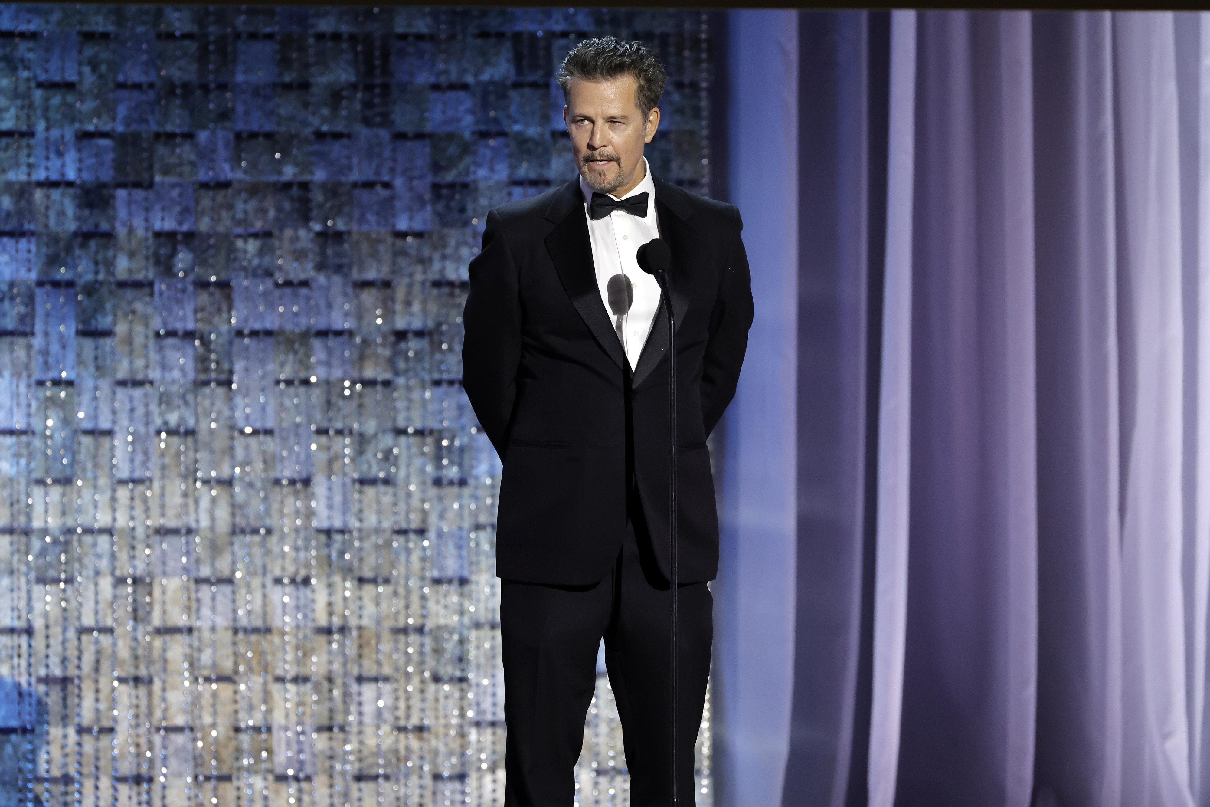 Ted King is seen presenting on stage at the 2022 Daytime Emmy Awards, dressed in a sharp black tuxedo with a bow tie. Standing with his hands behind his back, he speaks into a microphone against a shimmering, curtain-lined backdrop, bringing poise and formality to the televised event.