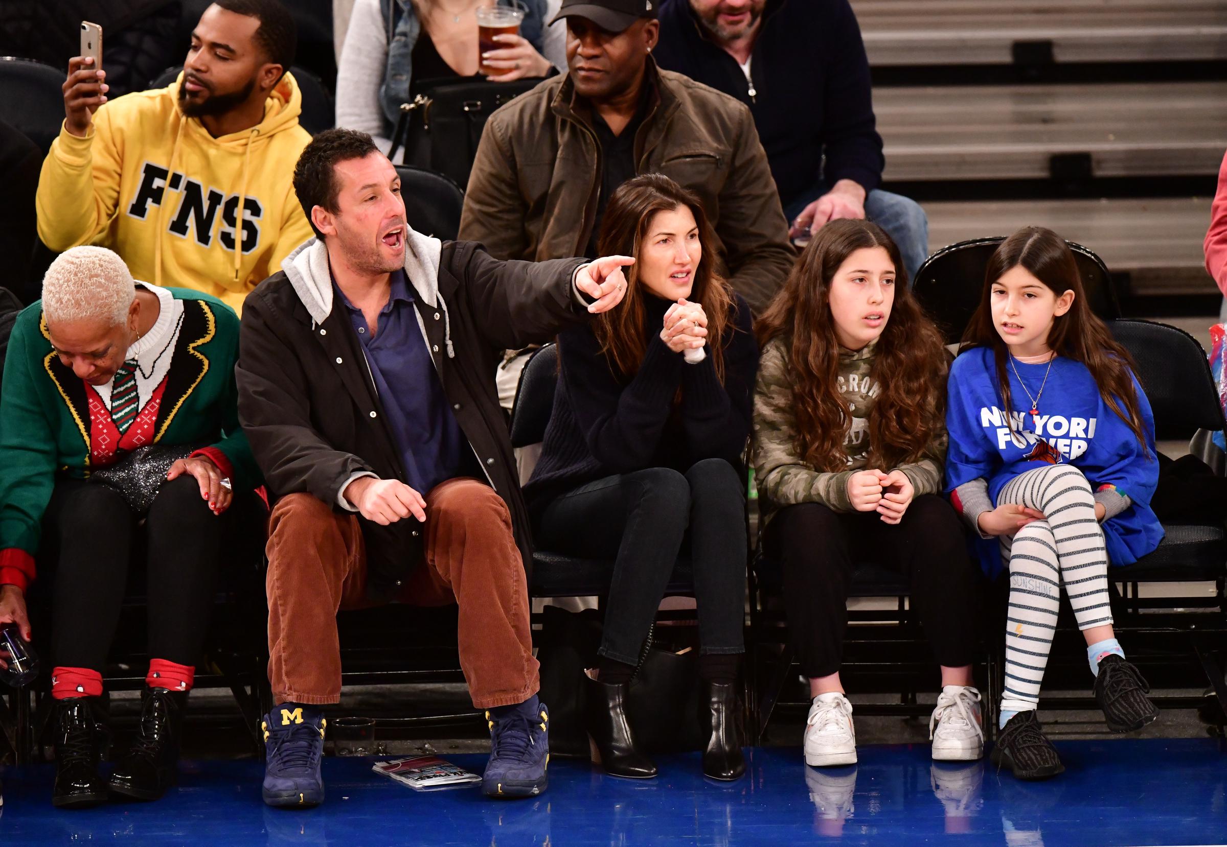 Adam, Jackie, Sadie, and Sunny Sandler traded Hollywood glamor for courtside fun in New York City as they attended the Milwaukee Bucks vs. New York Knicks game at Madison Square Garden. The family looked completely immersed in the action, with Adam animatedly pointing toward the court.