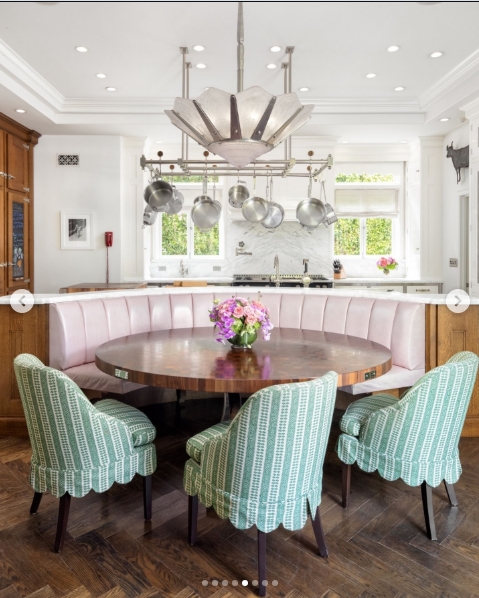 The kitchen eating area leans into colour in a way that is unmistakably Sharon's doing: a curved pink leather banquette wraps around a circular walnut table, with scallop-hemmed sage green chairs pulled up to meet it. A sculptural Art Deco chandelier hangs above a hanging pot rack, with marble splashback and garden views visible through the window beyond. | Source: Instagram/top.music.songs