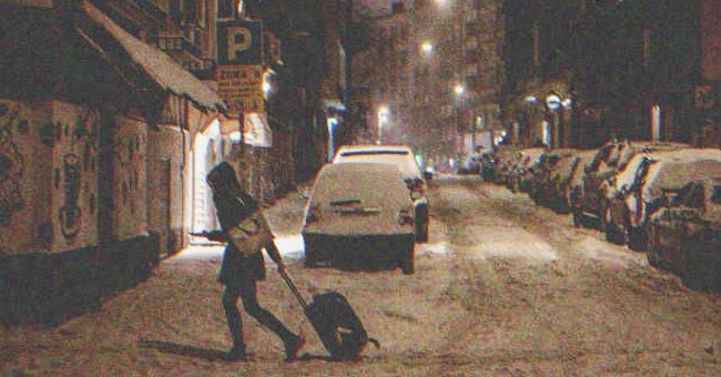 Mujer cruzando la calle de noche con una maleta. | Shutterstock