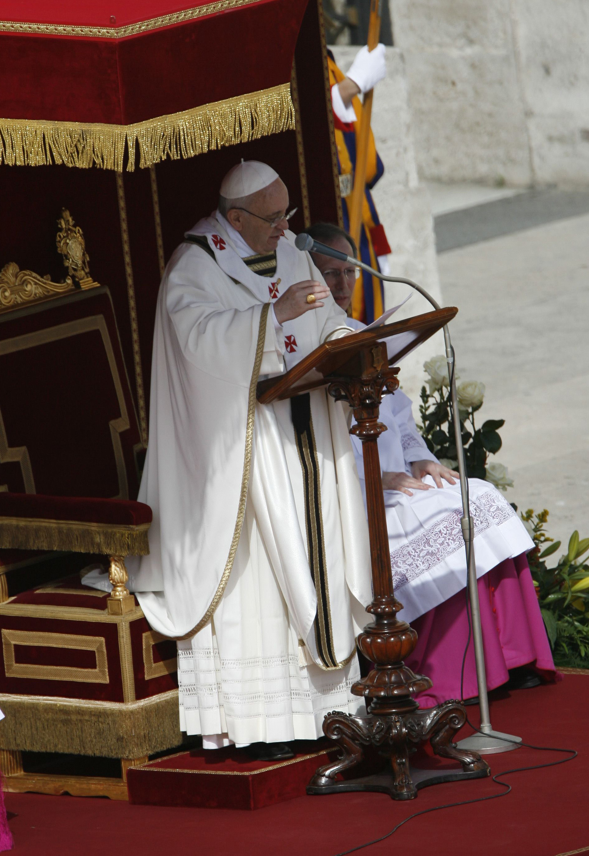Pope Francis delivers a sermon from a carved wooden lectern, draped in cream vestments with red crosses, standing beside a throne-like chair trimmed in gold