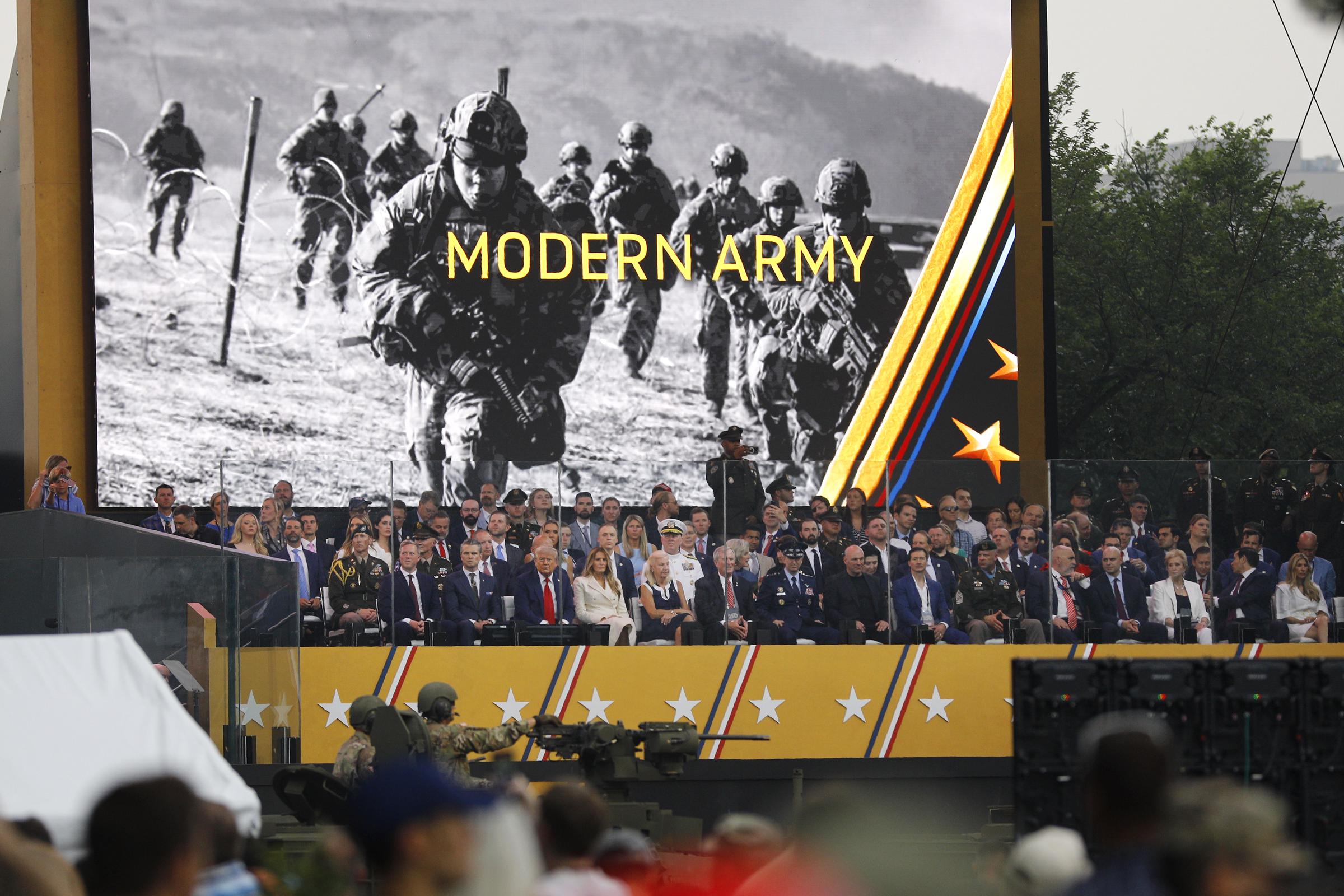 Donald and Melania Trump sit among dignitaries and military leaders, including Pete Hegseth and Army Secretary Daniel Driscoll, during the US Army's 250th anniversary parade. Behind them, a massive screen displays "MODERN ARMY" above combat imagery.