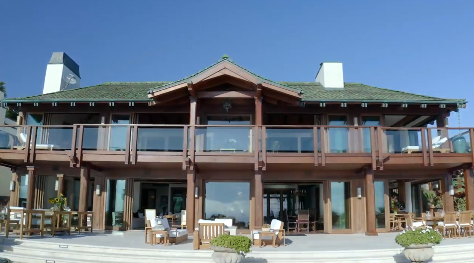 This ground-level view highlights the home's Thai-inspired architecture, featuring rich timber framing, large glass railings, and a green clay-tiled roof flanked by two white chimneys. Expansive sliding glass doors open the main living areas onto a sprawling outdoor terrace furnished with comfortable lounge seating and dining tables. | Source: YouTube/Architectural Digest