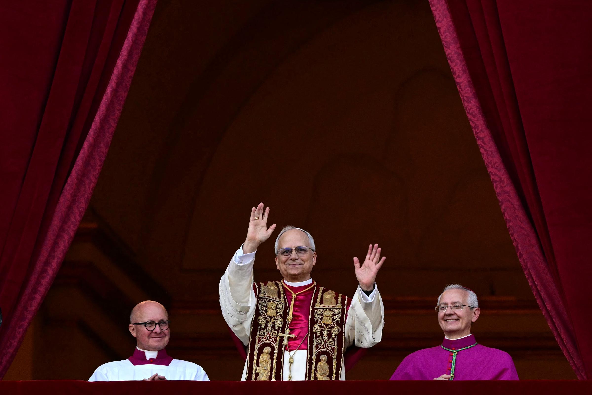 Pope Leo XIV raises both hands in greeting, wearing a richly embroidered stole over cream robes as he steps onto the balcony framed by deep red velvet drapes