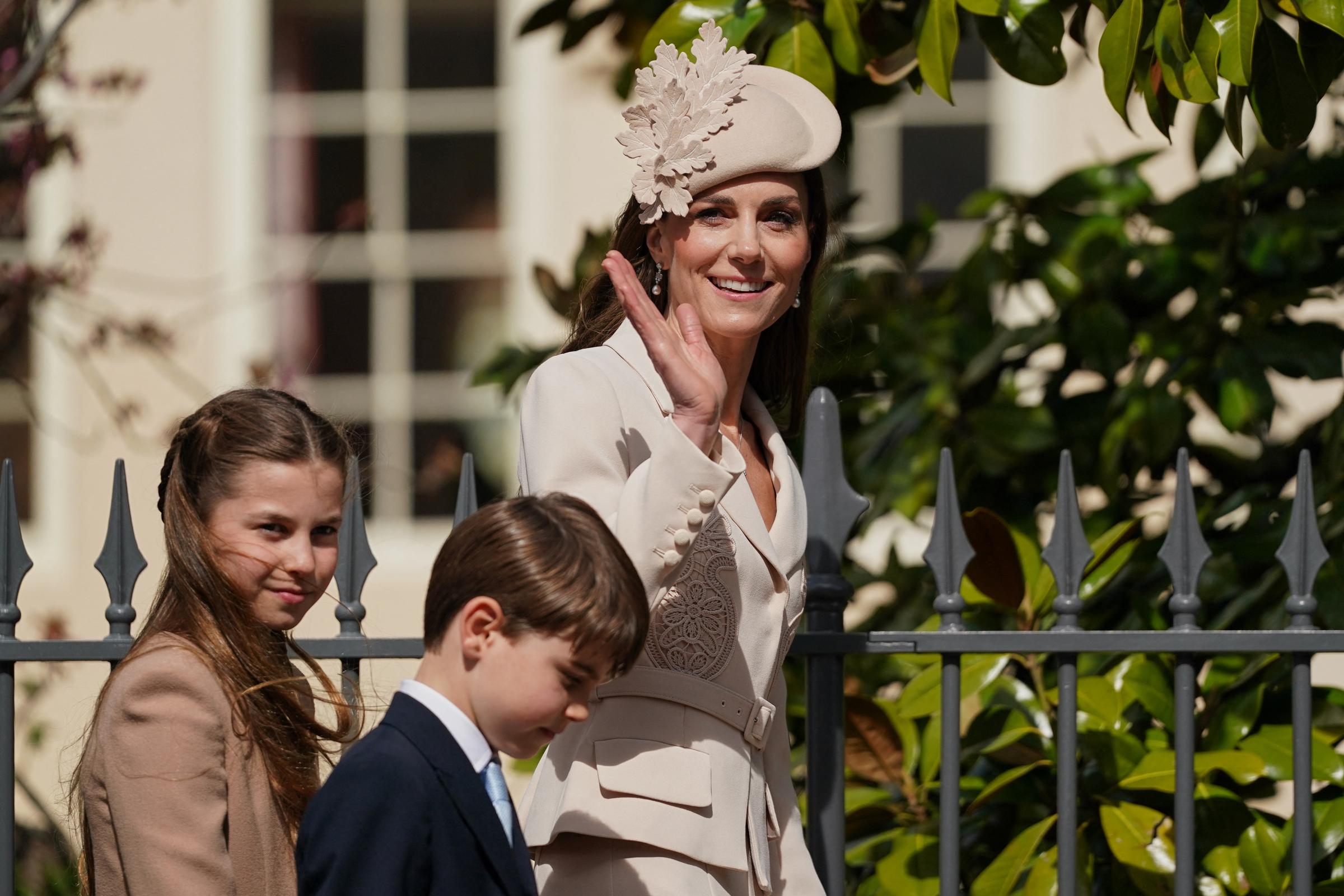 The Princess of Wales waves to the waiting crowds at Windsor, with Princess Charlotte and Prince Louis close beside her. Princess Charlotte, hair loose and windswept, catches the moment with a quiet sideways smile, while Prince Louis, head bowed in his navy suit and pale blue tie, appears entirely unbothered by the assembled well-wishers.