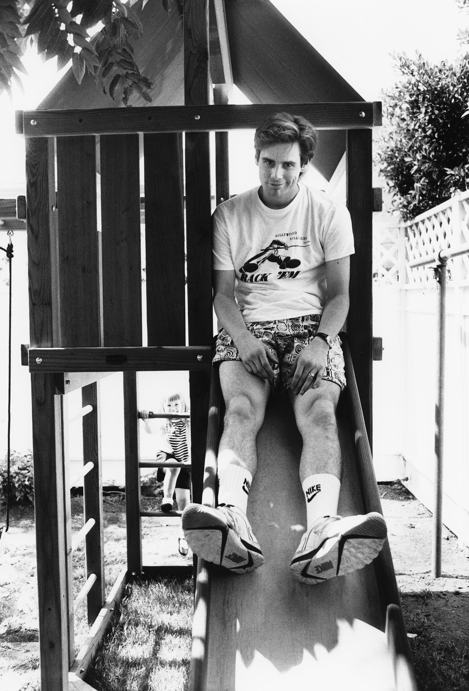 In this charming black-and-white moment, Jim Carrey sits at the top of a backyard slide, sneakers outstretched and signature smirk in place. Behind him, his young daughter Jane climbs up the play structure.