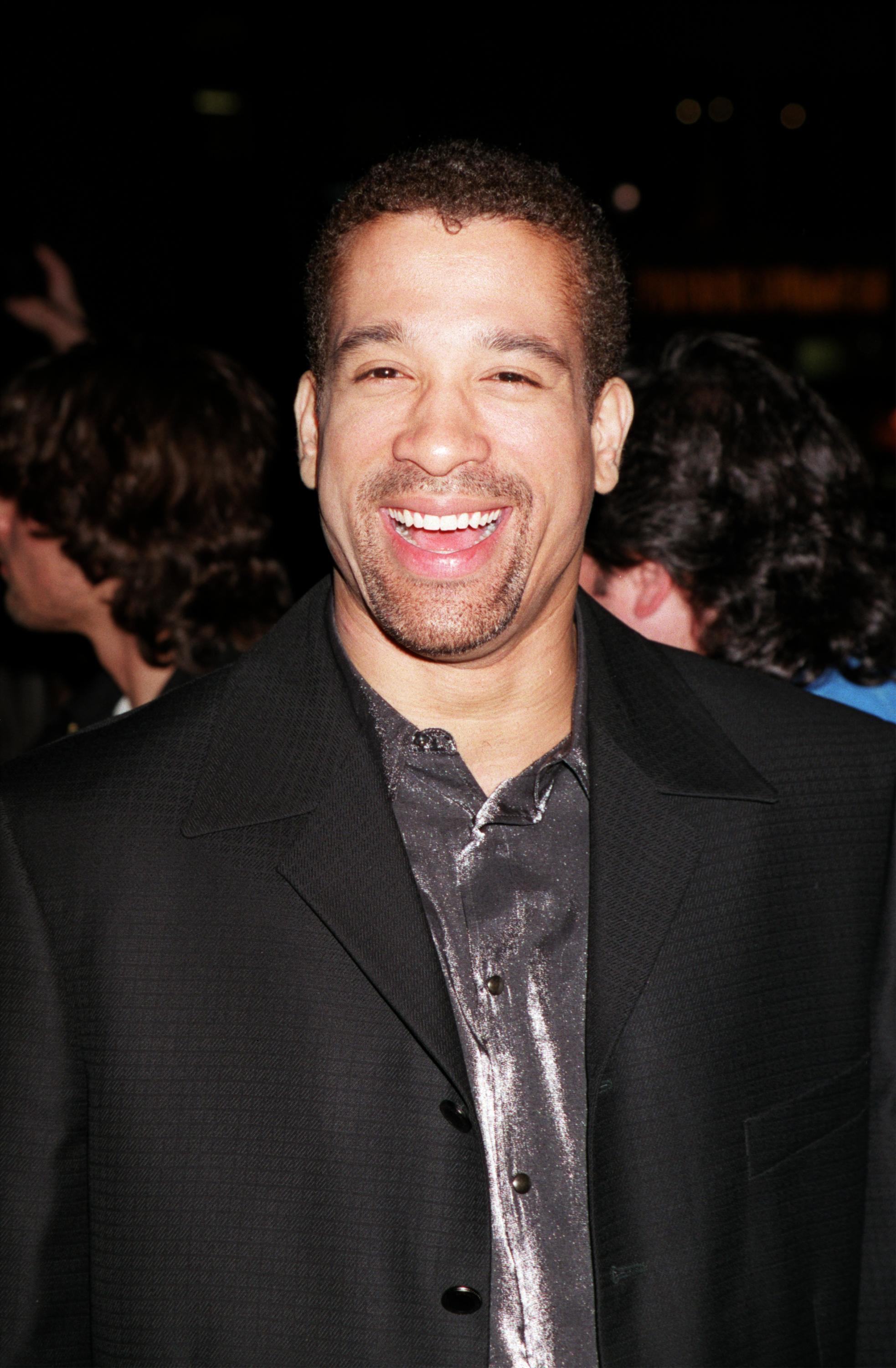Dorian Gregory is seen at a red carpet event in the early 2000s, smiling brightly in a dark suit over a metallic silver shirt. The nighttime setting and crowd in the background suggest a lively atmosphere, while his relaxed and cheerful demeanor adds to the candid charm of the moment.