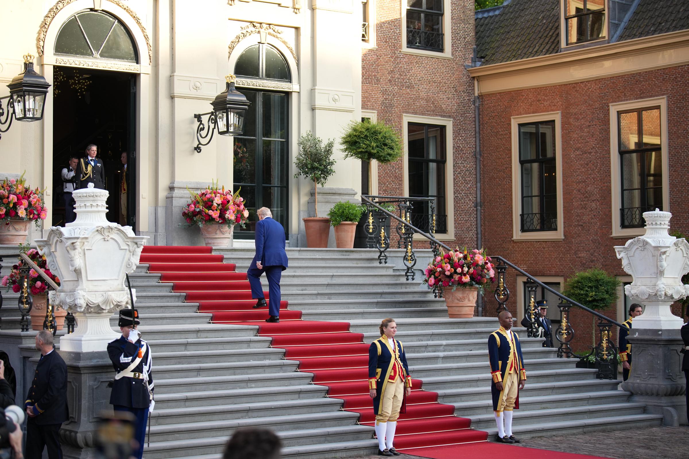 The grandeur of the residence is on full display as Trump ascends the long red-carpeted staircase, flanked by two royal guards standing at attention. Elegant floral arrangements line either side, enhancing the regal setting as he makes his way toward the entrance of the palace.