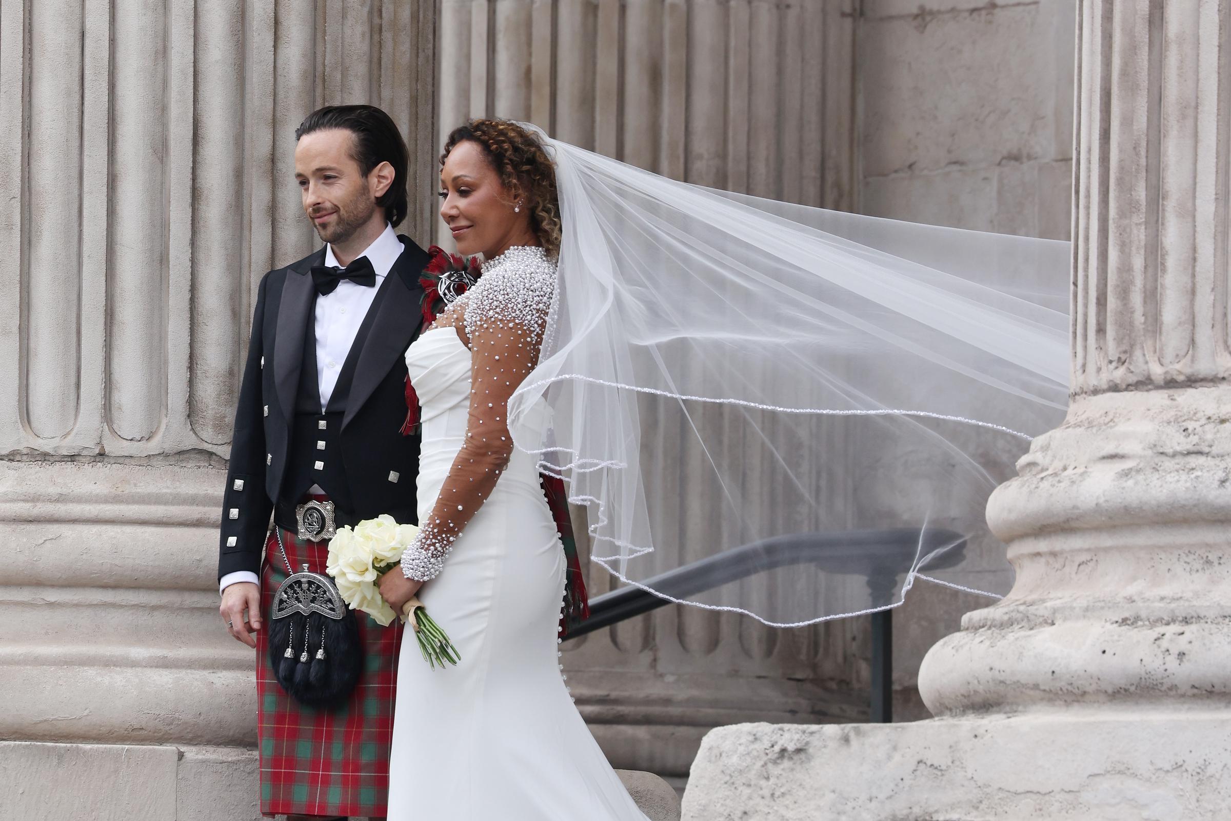 In a medium shot taken outside St. Paul’s Cathedral, newlyweds Rory McPhee and Mel B stand side by side. The grand backdrop highlights the significance of the moment as the couple pauses, smiling softly after exchanging vows.