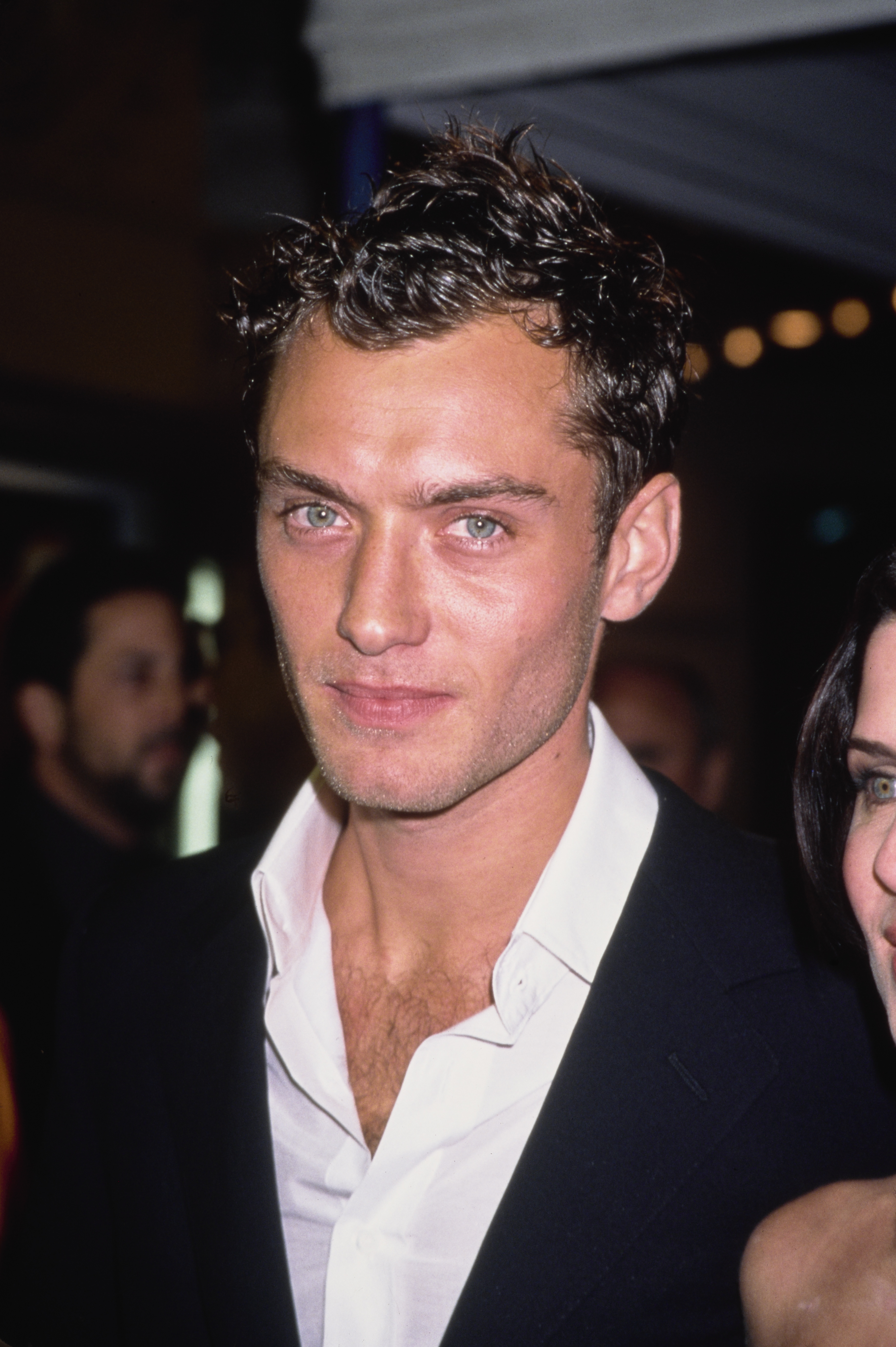 A close-up of a 20-something Jude Law at the Vanity Fair Oscar Party in West Hollywood, March 2000 — sporting his dark, tightly curled hair, pale blue-green eyes, and an open-collar white shirt beneath a black blazer.