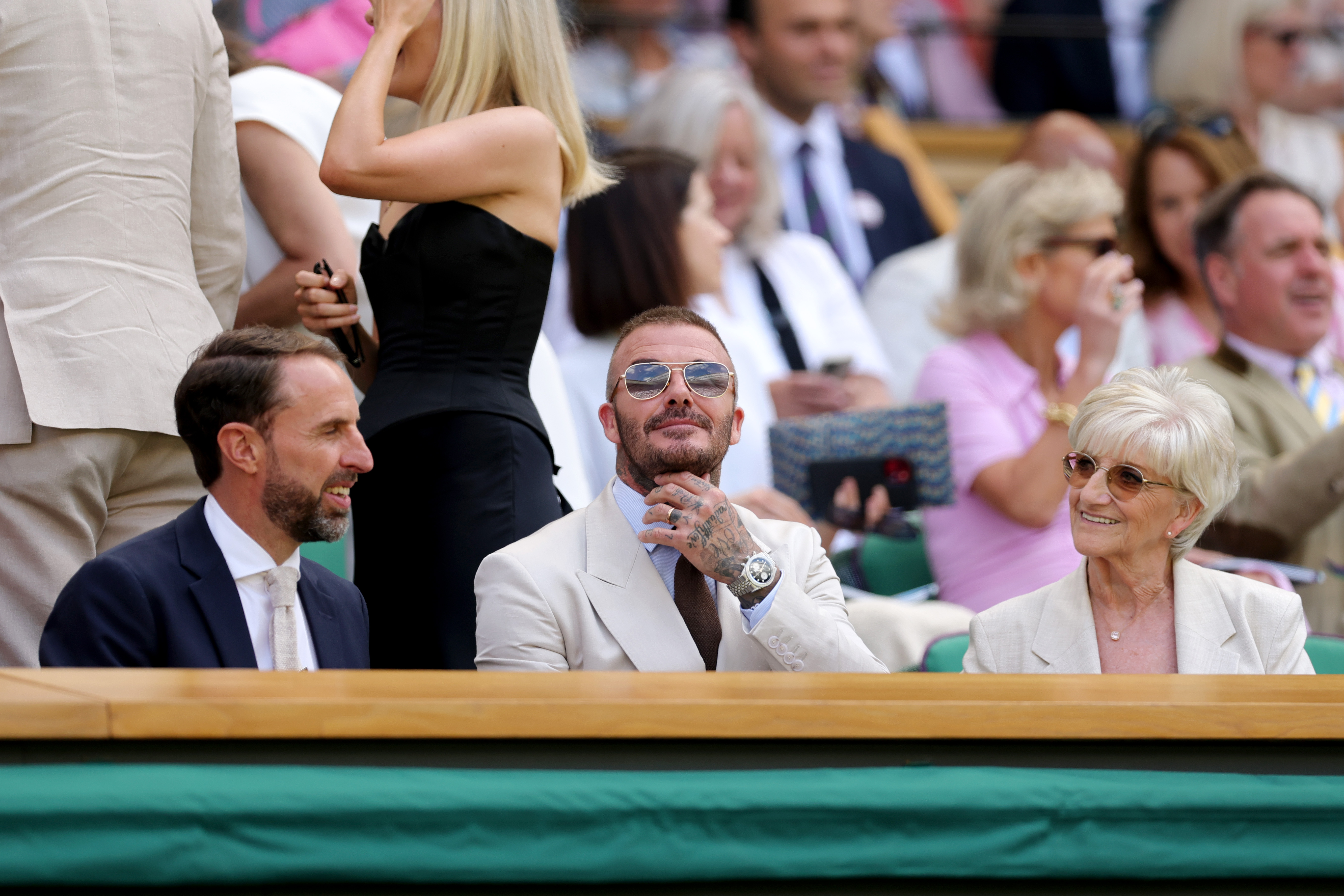 David Beckham sits between Gareth Southgate and his mother, Sandra Georgina West, at Wimbledon. With his hand resting on his chin, he appears thoughtful as he takes in the action from the Royal Box.