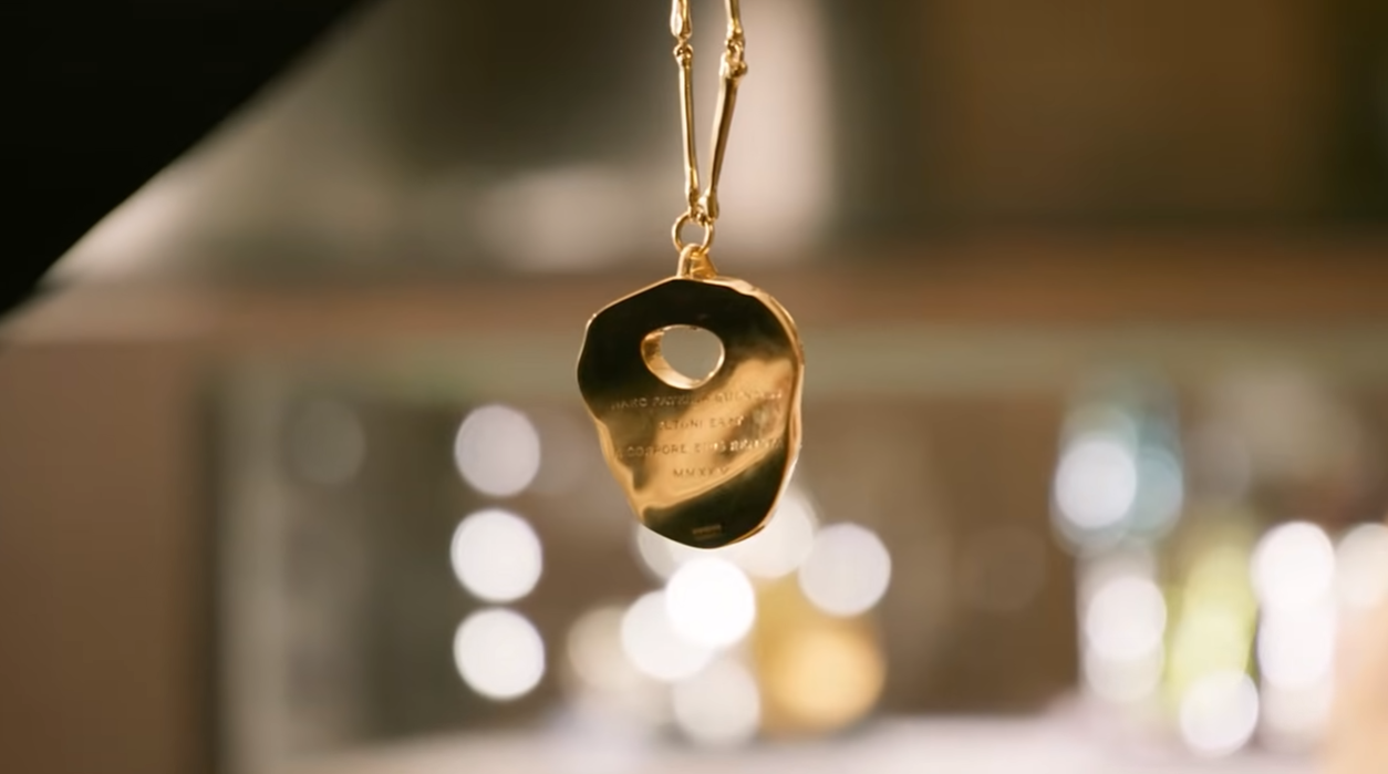 The back of the kneecap necklace, fully gilded in gold, twists on its delicate bone-link chain to reveal the Latin inscription engraved across its uneven surface — catching the light against a soft, bokeh backdrop. | Source: YouTube/World Gold Council