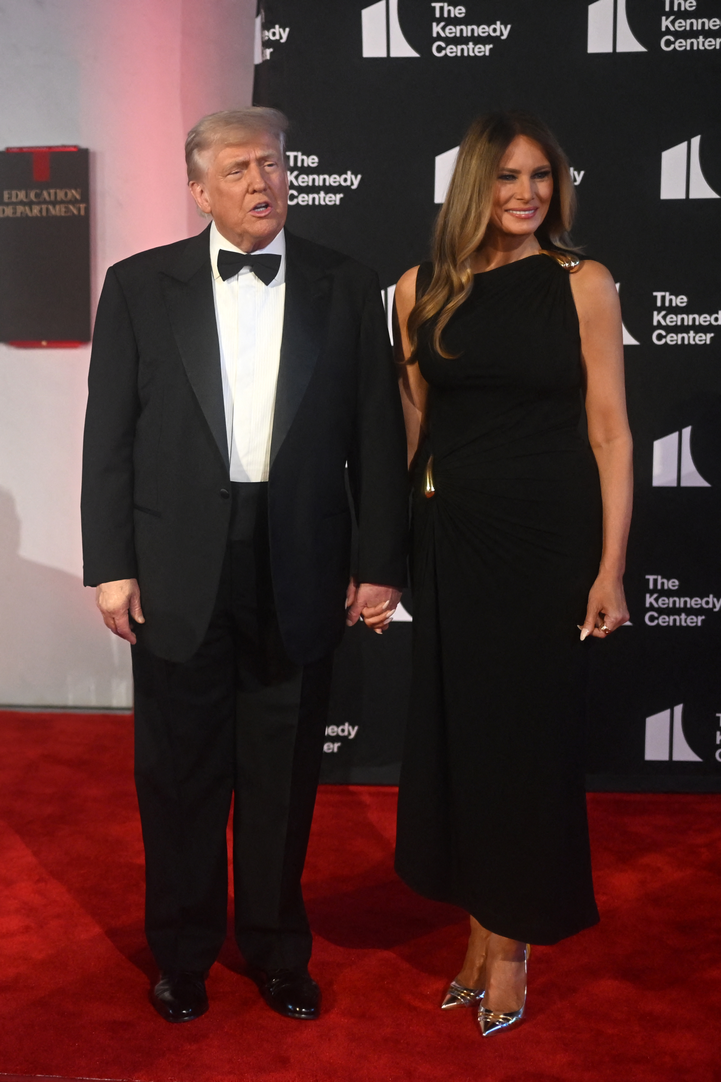 On the red carpet, Donald and Melania stand close, holding hands. Melania’s full look is once again visible—her black dress cinched at the hip with gold detailing, paired with gleaming metallic heels. She smiles warmly, her hair styled in soft waves, while Donald appears mid-sentence, adding a candid touch to the formal moment.