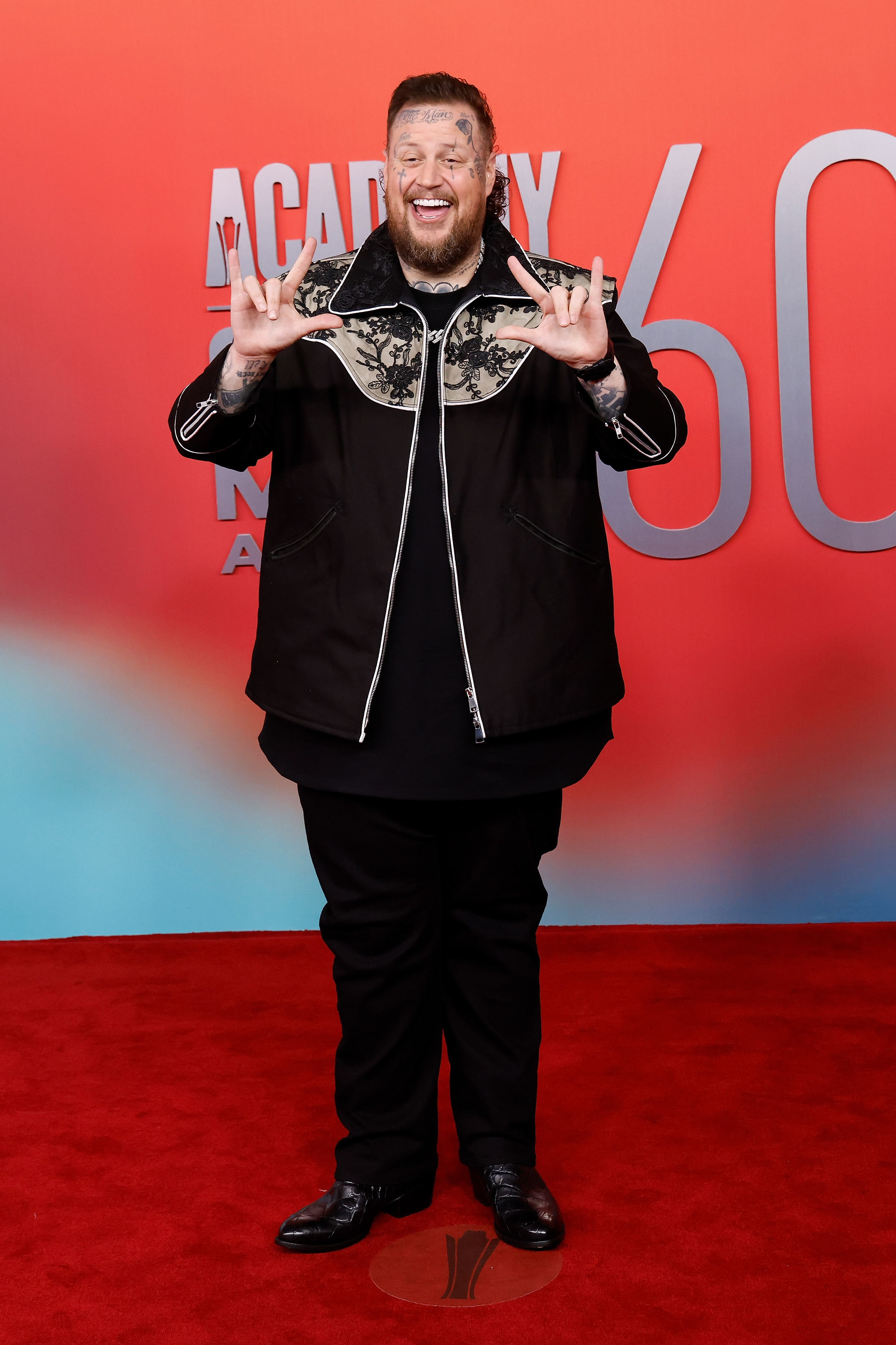 Jelly Roll attends the 2025 ACM Awards in Frisco, Texas, bringing big personality and bigger energy to the red carpet. The artist wears a two-tone zip-up jacket and flashes signs with his hands, radiating excitement ahead of the show.