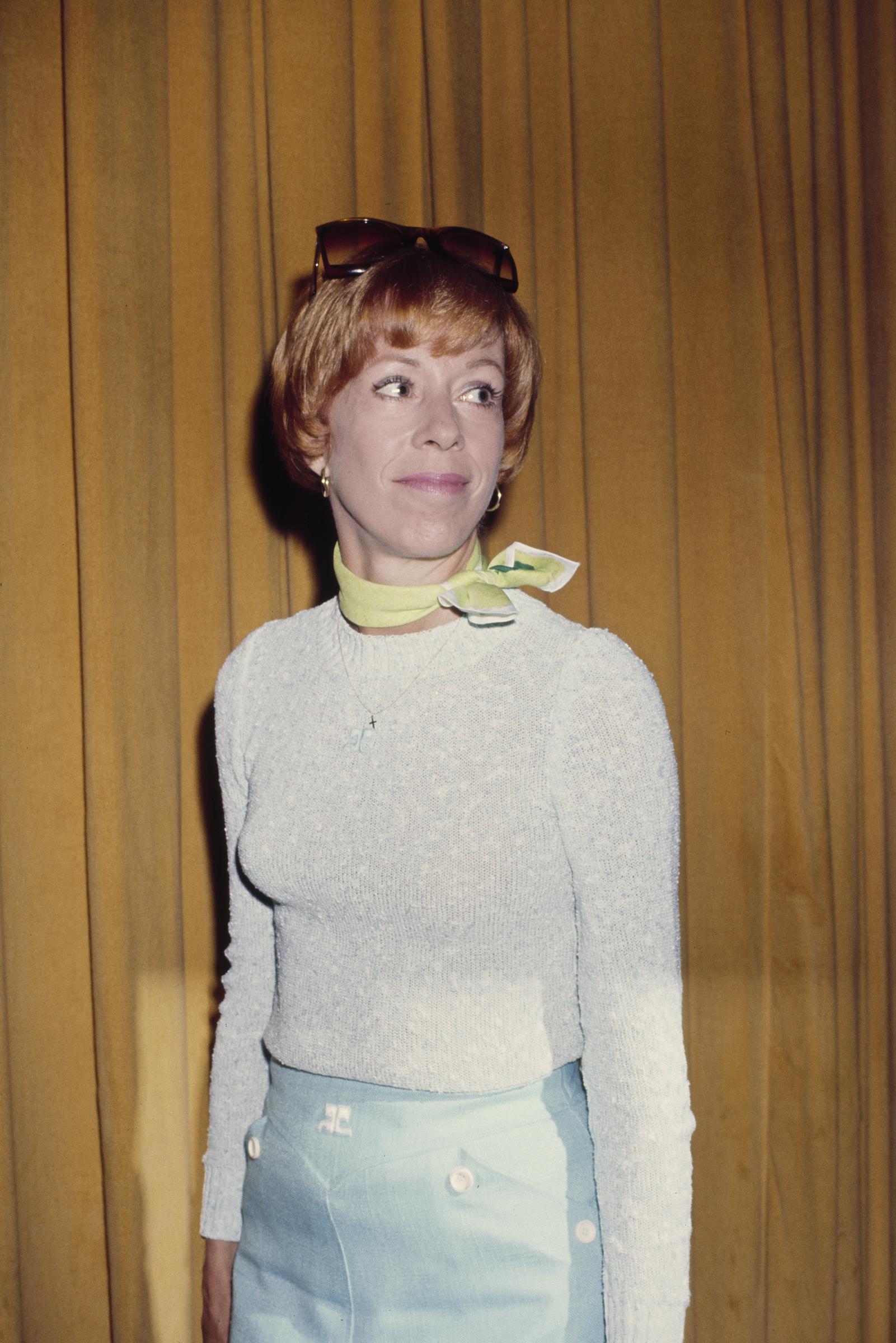 She pairs a light grey crew neck sweater with a high-waisted blue skirt, accessorized with a light green neckerchief tied at the side of her neck. Sunglasses rest on her head as she looks off to her left.