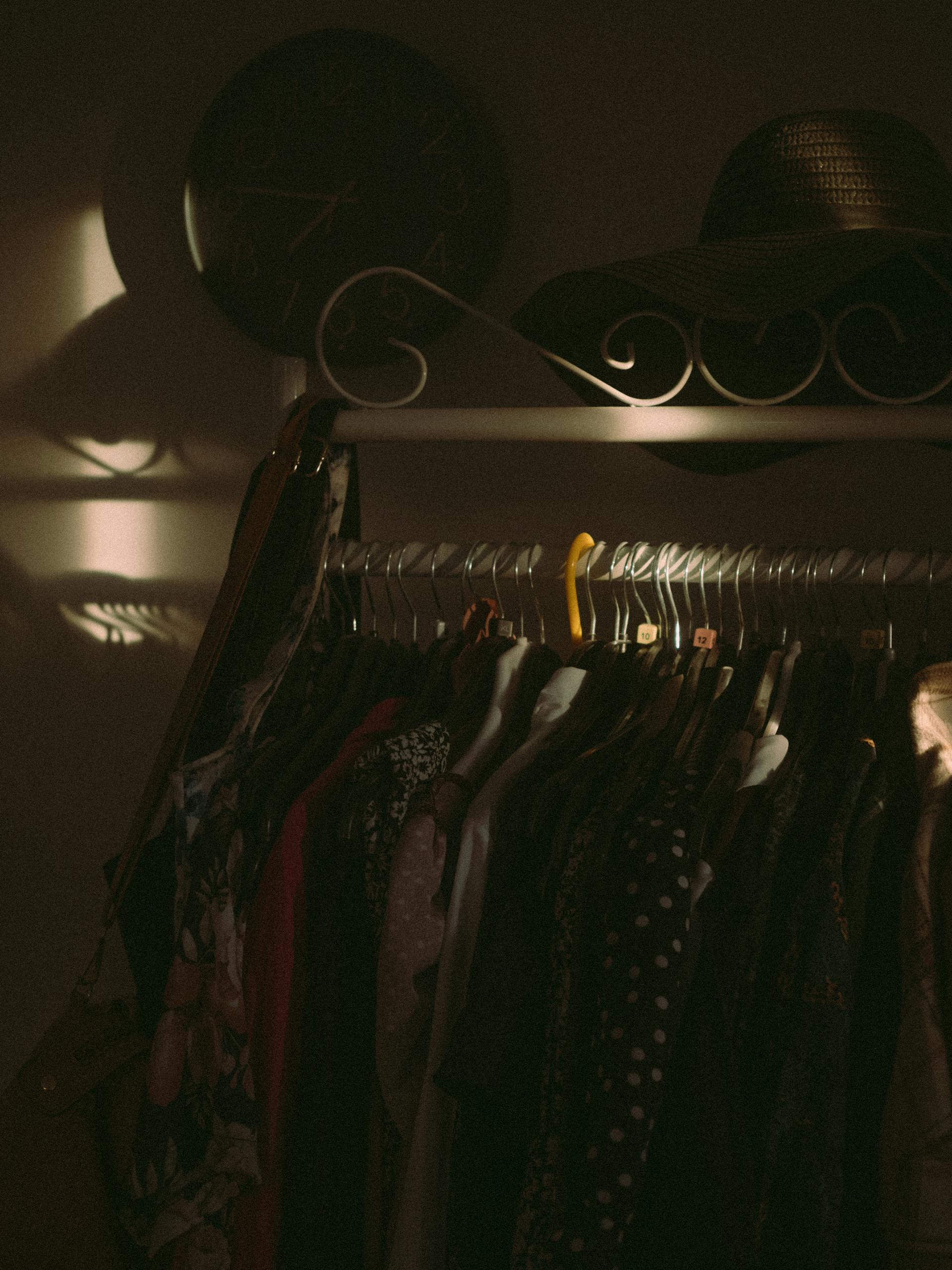 A dark shot of clothes hanging on a rack with a hat on top and a clock hanging on the wall | Source: Pexels