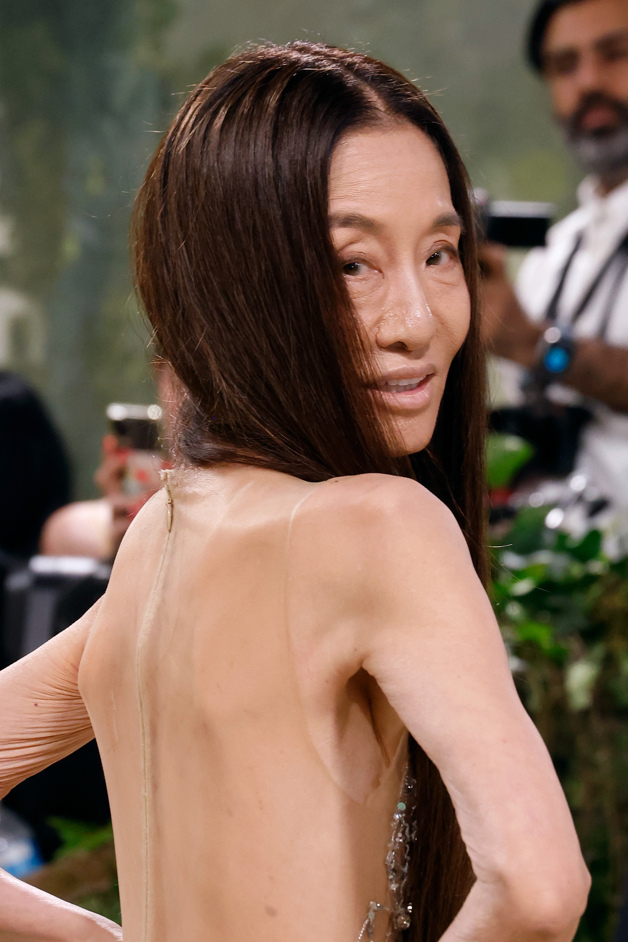 Vera Wang turns toward cameras with a soft smile at the Met Gala, wearing a sheer, embellished gown with a fully open back. Her long, straight hair is swept away from her back, allowing the delicate detailing of the dress to take center stage.
