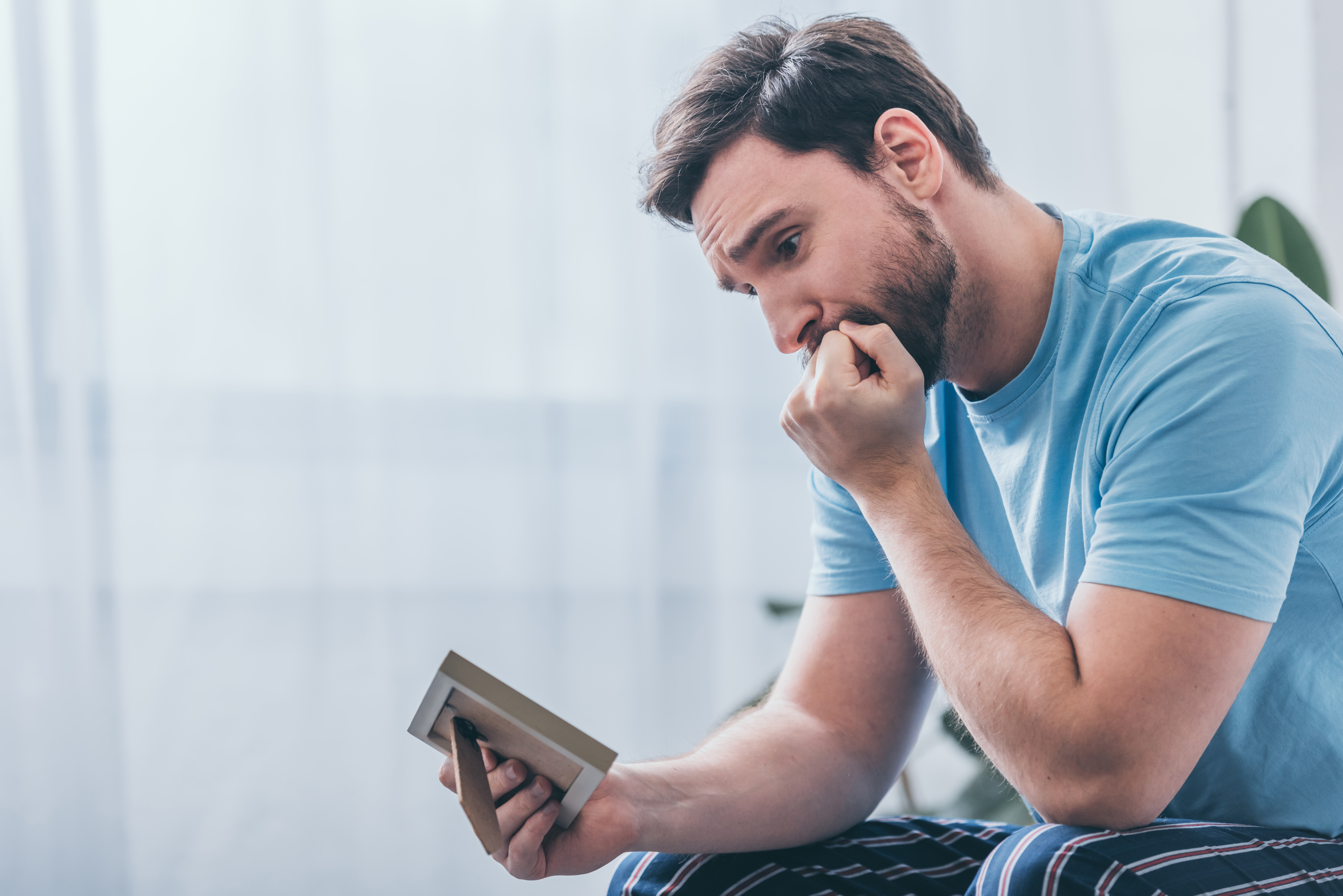 depressed man sitting, covering mouth with hand and looking at photo frame at home | Source: Shutterstock