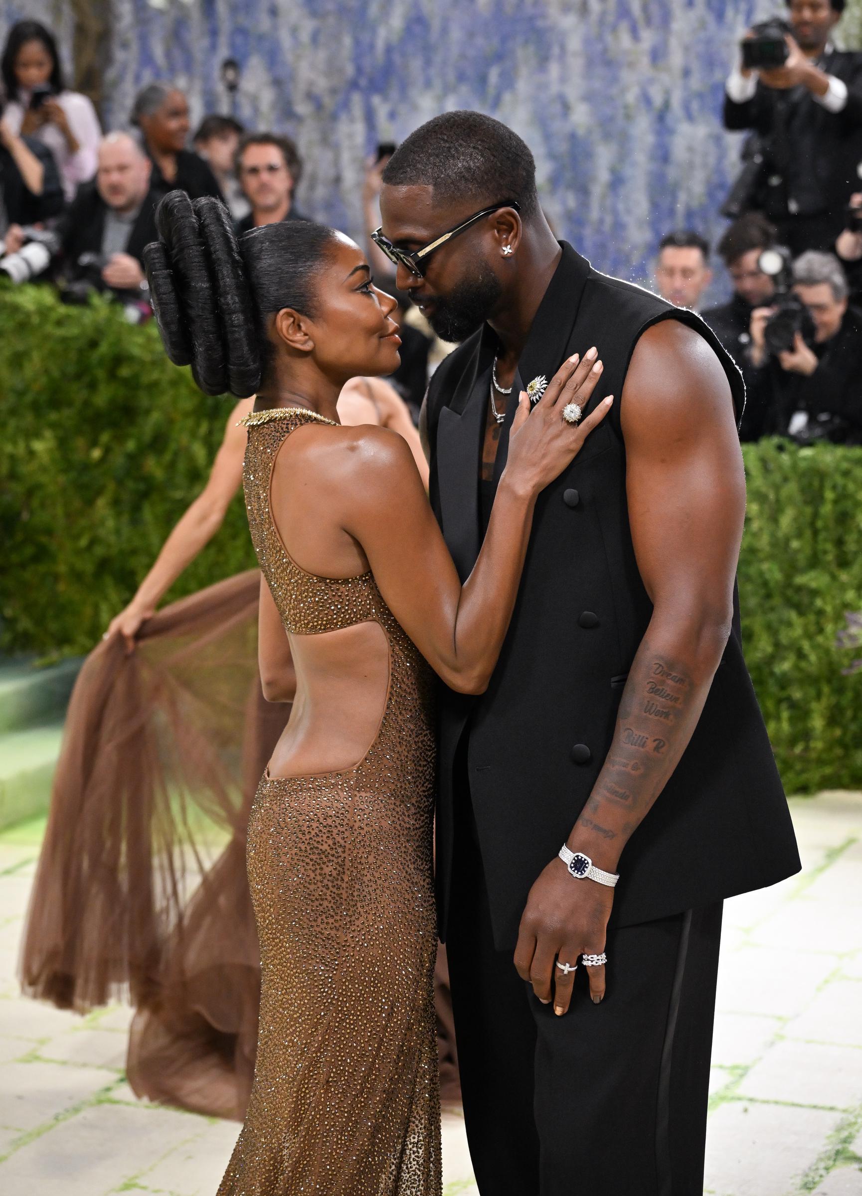 Gabrielle Union stuns in a shimmering bronze gown with a sleek, sculpted silhouette and statement necklace. Facing her, Dwyane Wade leans in wearing a sleeveless black suit, their close pose capturing an intimate, stylish moment.