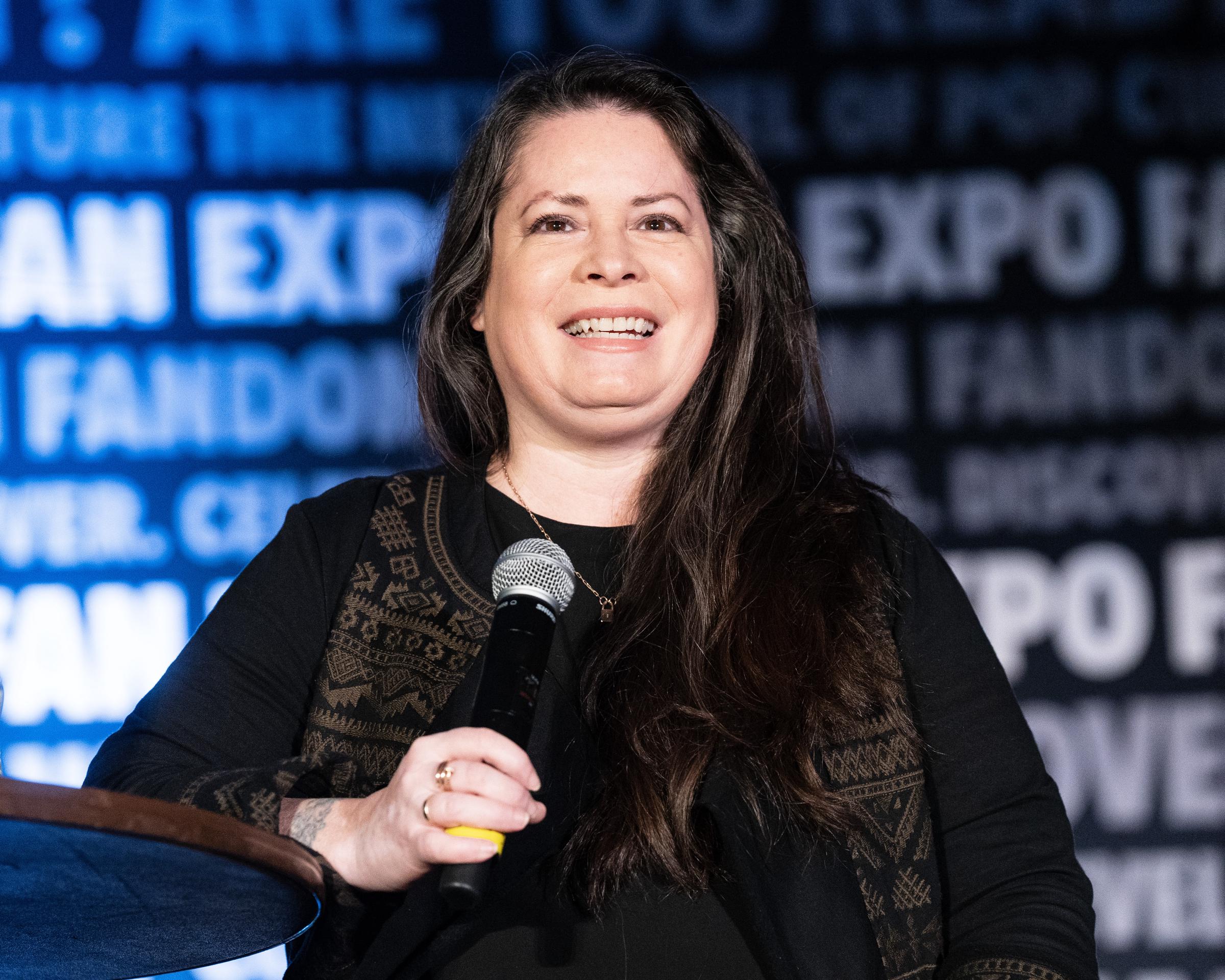 Holly Marie Combs is seen speaking at FAN EXPO Denver in 2024, holding a microphone and smiling brightly. Wearing a black outfit with a patterned vest, she stands confidently against a backdrop filled with event branding, projecting warmth and ease during the fan convention appearance.