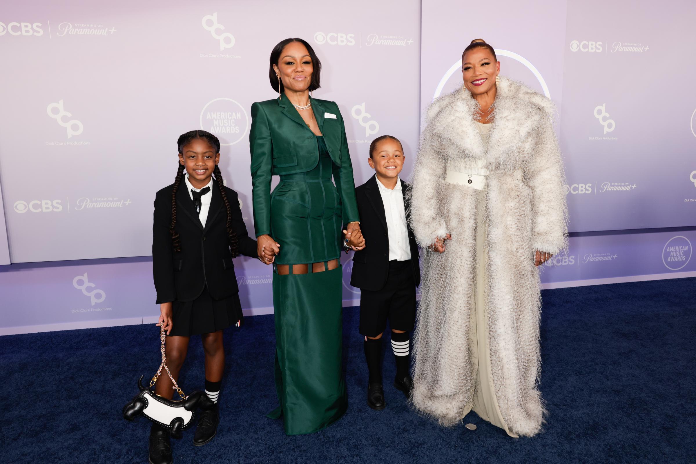 Eboni Nichols and Queen Latifah pose with two children on the blue carpet, creating a coordinated family moment beneath the glowing AMAs backdrop.