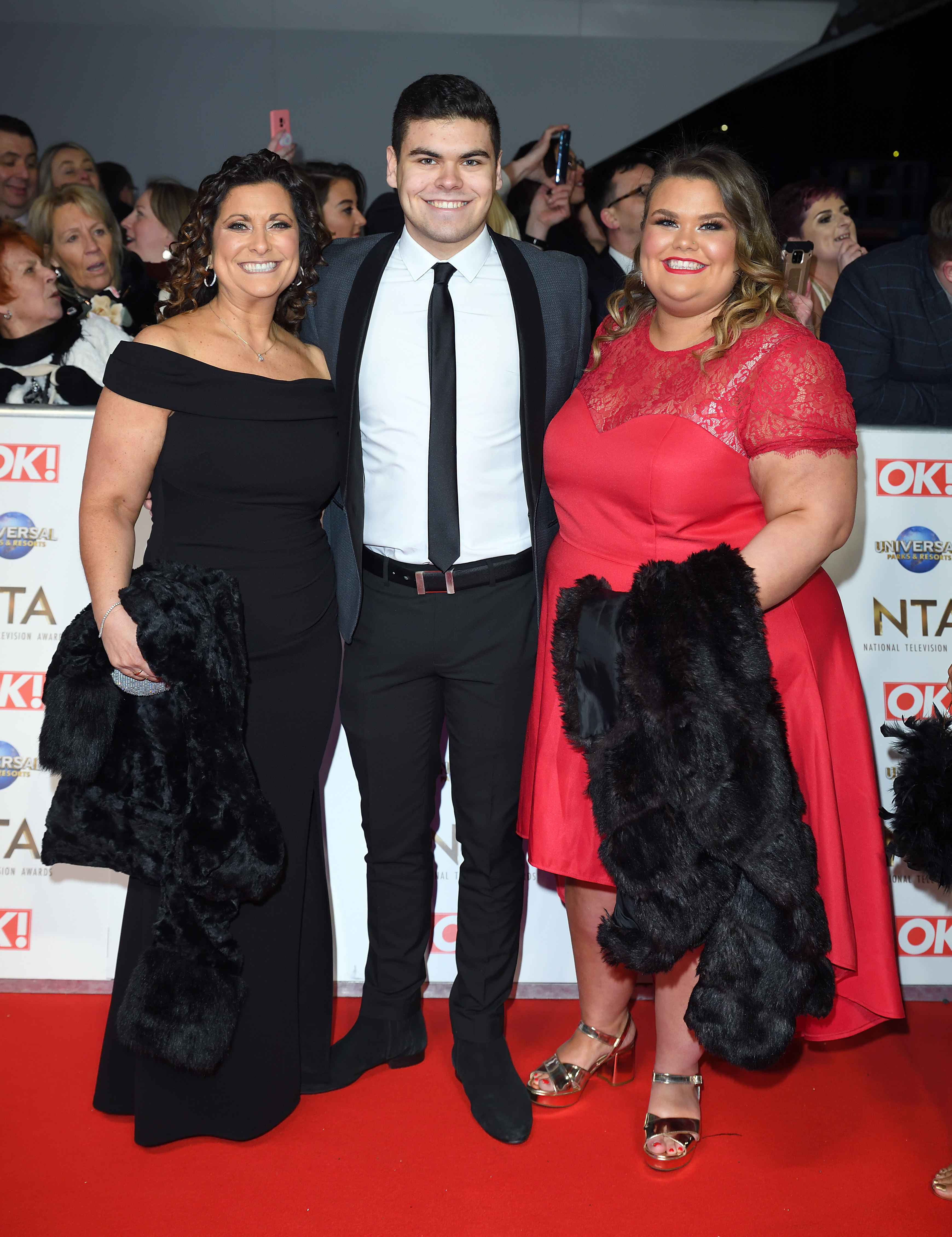 Tapper, in a red lace cap-sleeve dress and gold strappy heels, arrives with her mother Nikki — in an off-shoulder black gown — and her suited brother Josh on the NTA red carpet at The O2 Arena, London, January 2020. All three are grinning broadly as fans and photographers crowd behind them.