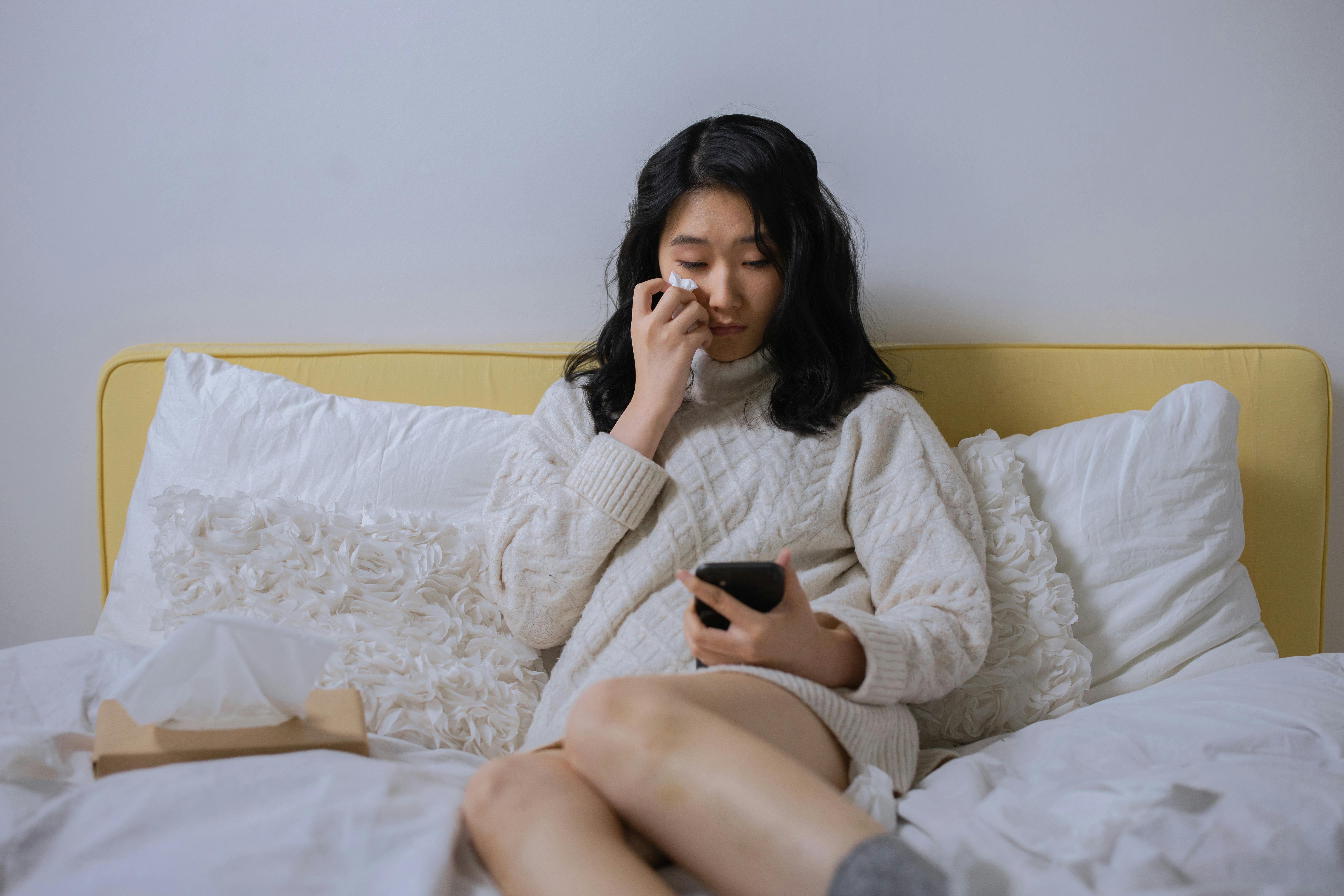 A woman crying and wiping tears while holding a phone in bed | Source: Pexels