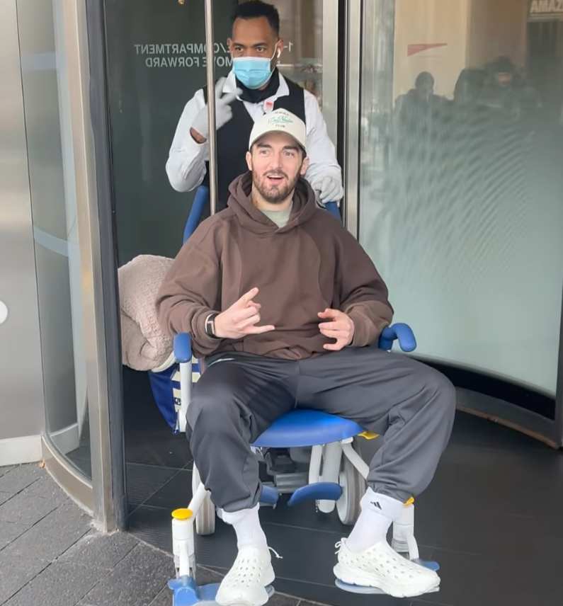 Daniel Neeson sits in a wheelchair outside the hospital as a masked staff member pushes him through the doors. | Source: Instagram/bgdans91