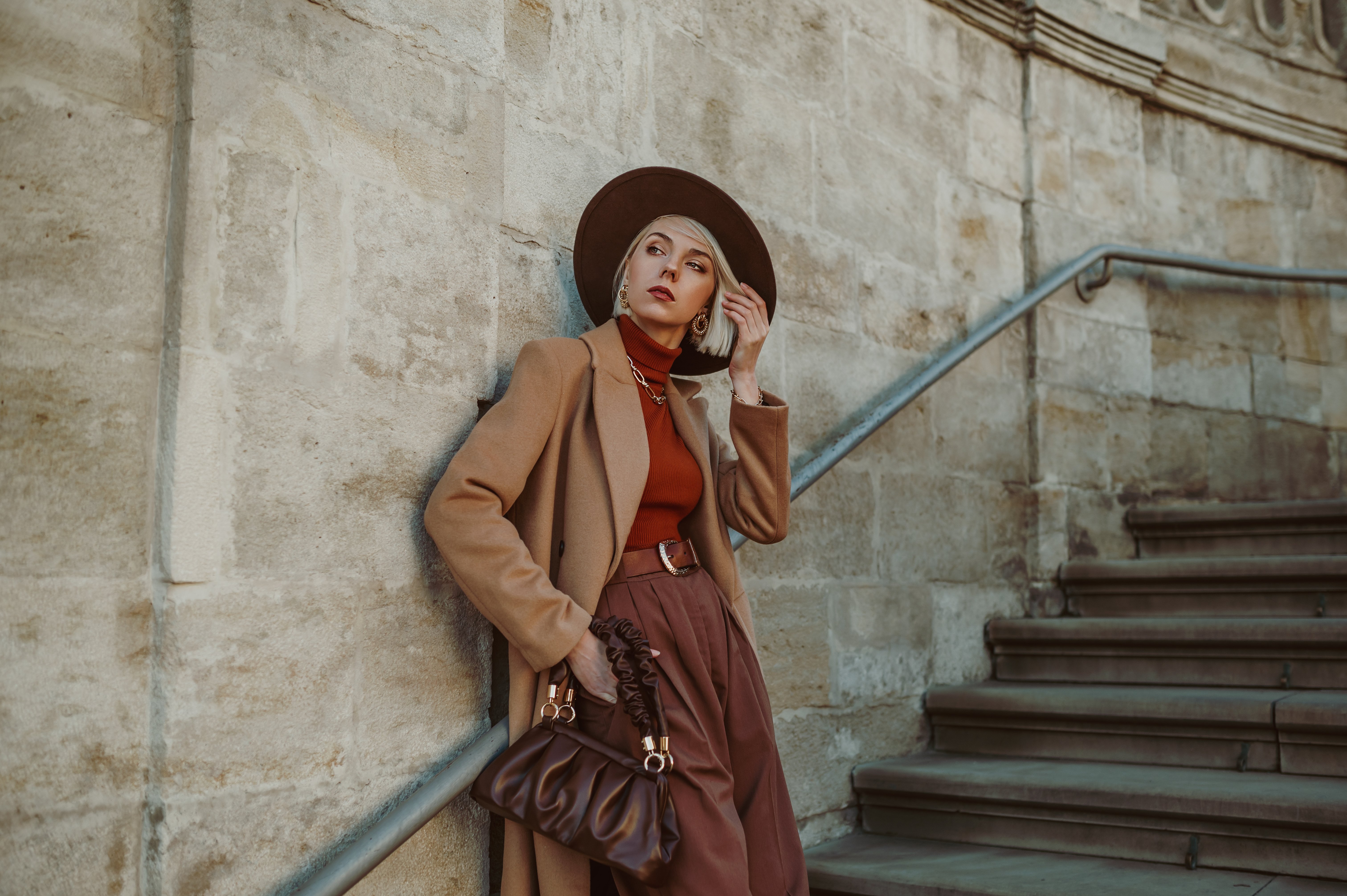 Une femme élégante et luxueuse vêtue d'un beige midi branché, d'un manteau à chameaux, d'un chapeau marron, d'un pantalon, d'un col de fer orange, tenant un sac main en cuir, posant dans la rue de la ville - Photo : Shutterstock