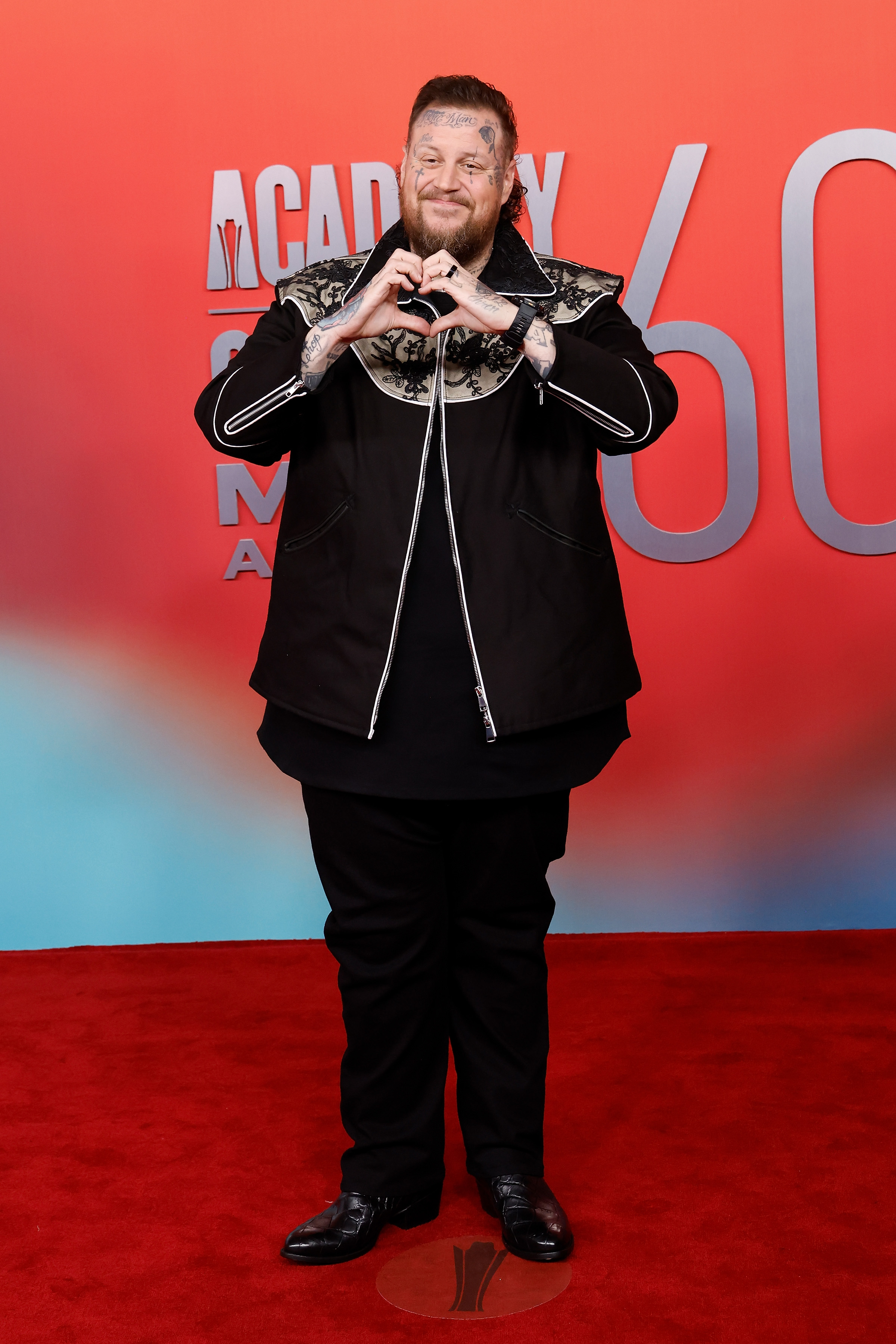 Jelly Roll attends the 2025 ACM Awards at Omni Frisco Hotel in Frisco, Texas. He poses playfully on the red carpet, forming a heart with his hands while dressed in a black zip-front jacket with a patterned yoke and matching pants.