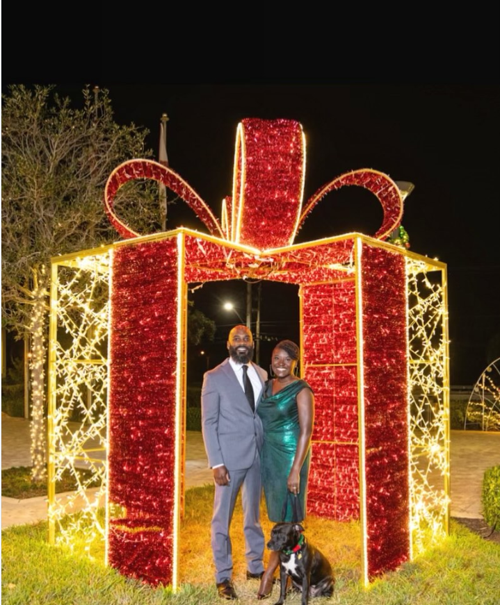 Nancy Metayer Bowen stands beside her husband, Stephen Bowen, as they pose under a large, illuminated holiday display shaped like a wrapped gift, both dressed in formal attire, with their dog sitting calmly at their feet amid the festive lights. | Source: Instagram/kingsb85
