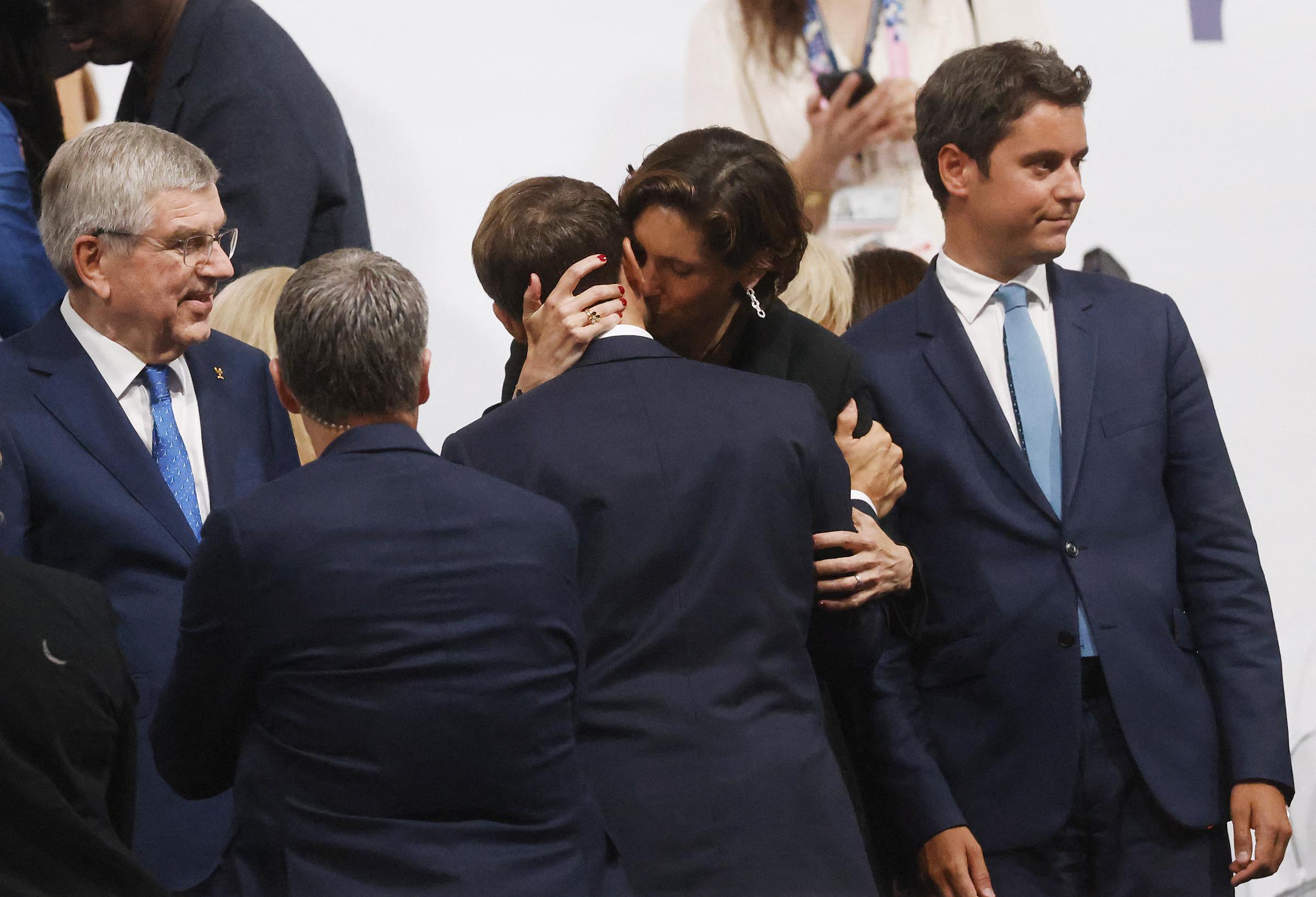 Amélie Oudéa-Castéra embraces Emmanuel Macron with both arms, placing one hand on the back of his head while drawing him in closely. Her face touches his neck as several individuals, including Thomas Bach, stand in view.