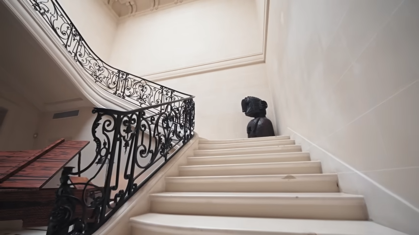 Looking up the soaring staircase toward a striking black sculpture that anchors the middle landing of the entryway. | Source: YouTube/@Archdigest