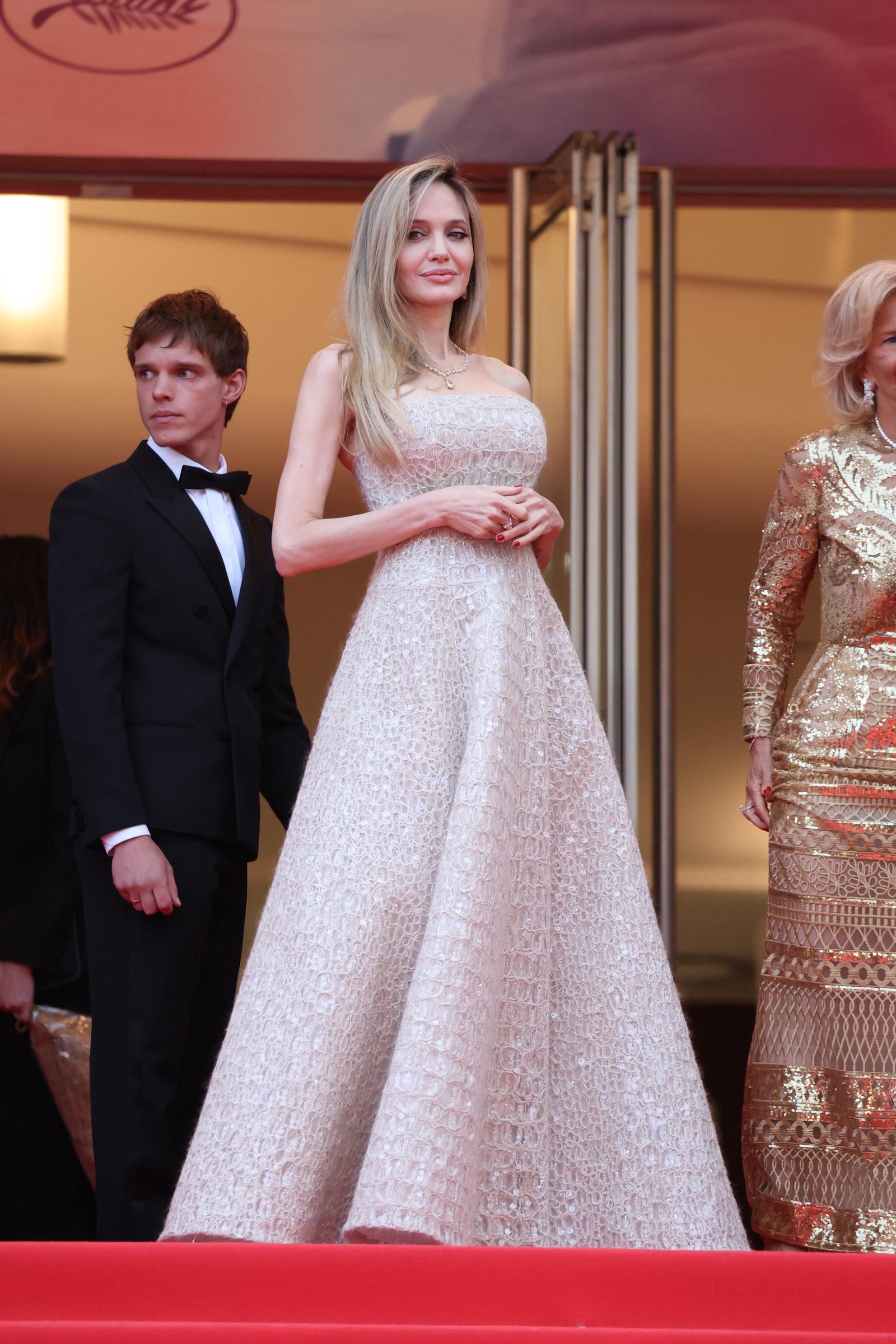 Angelina Jolie stands tall at the top of the Palais des Festivals staircase, hands gently clasped in front of her intricately detailed Brunello Cucinelli gown, exuding timeless poise against a backdrop of red and gold.