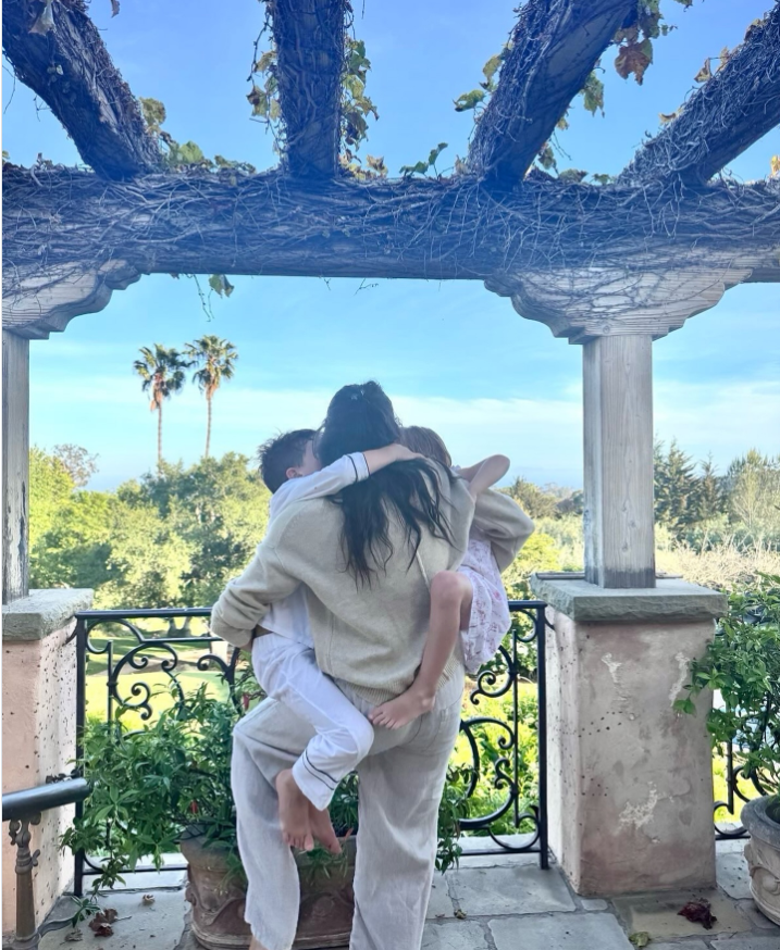 Meghan Markle stands beneath a vine-covered pergola while embracing Prince Archie and Princess Lilibet in a joyful family hug. | Source: Instagram/meghan