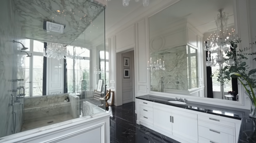 A long white double vanity with sleek black marble countertops faces a grand wall mirror, reflecting the room's high-end, light-filled design. | Source: YouTube/@Archdigest