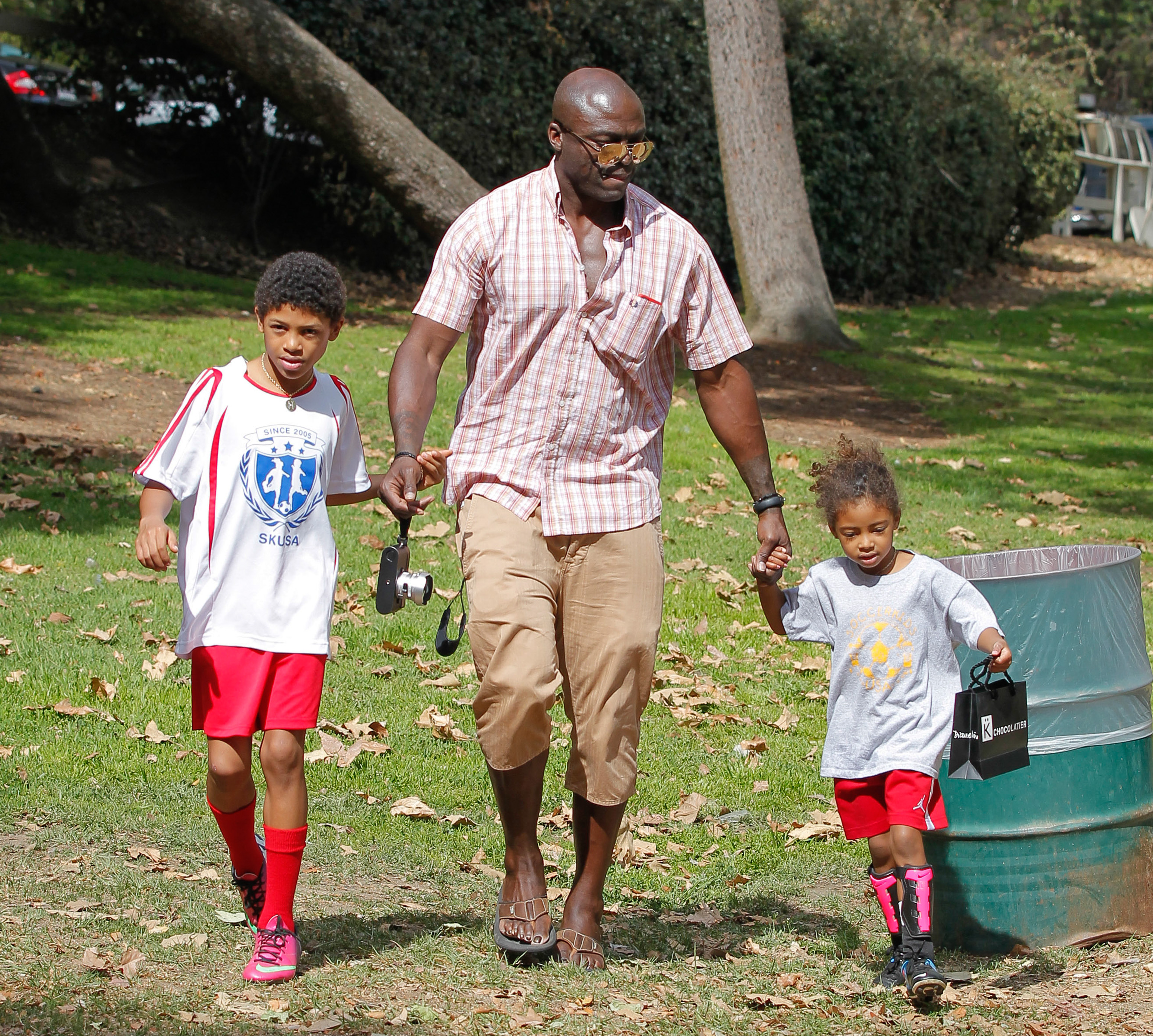 Under the dappled sunlight of a park in Los Angeles, Seal walks hand in hand with Lou Samuel, while Henry Samuel keeps pace beside them — an easy, grounded family moment where nature, movement, and quiet connection come together.