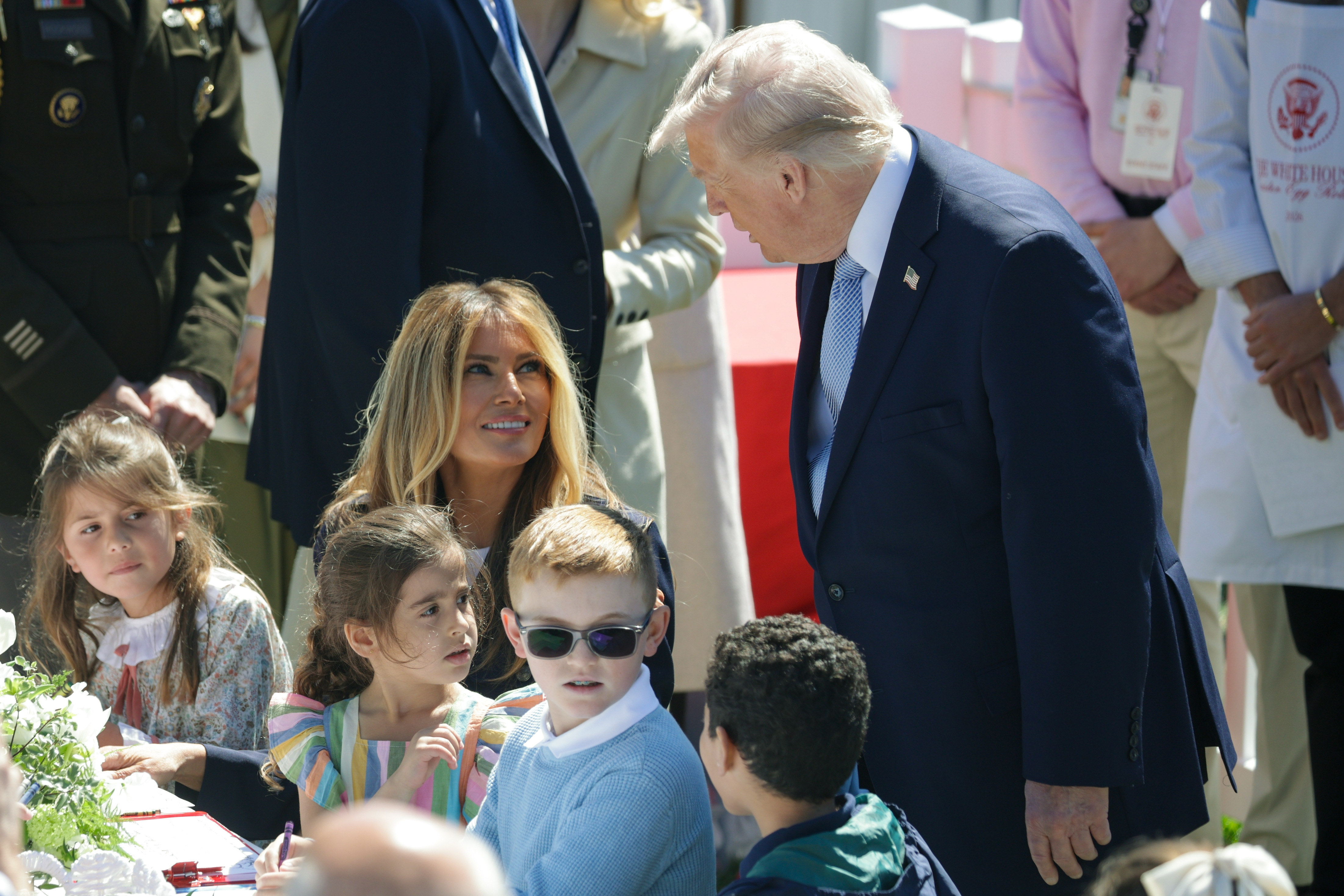 Donald Trump leans in to speak with Melania Trump and seated children as Melania looks up at him during the 2026 White House Easter Egg Roll, capturing a quieter moment from the South Lawn festivities.