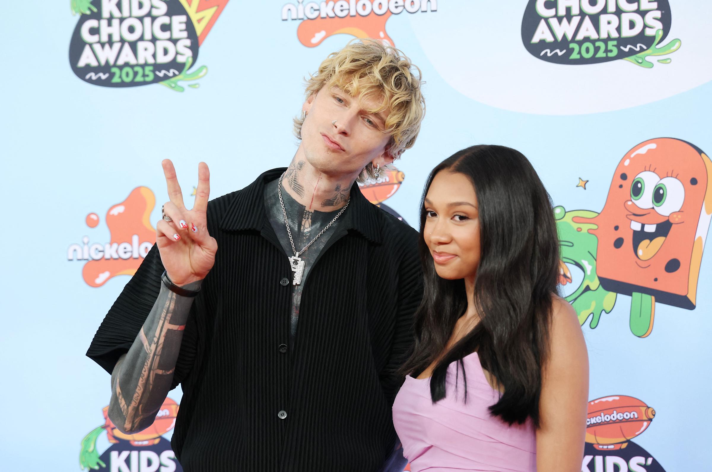 Machine Gun Kelly throws up a peace sign while Casie Baker keeps it cool beside him. Their laid-back pose and casual charm stand out on the 2025 orange carpet.