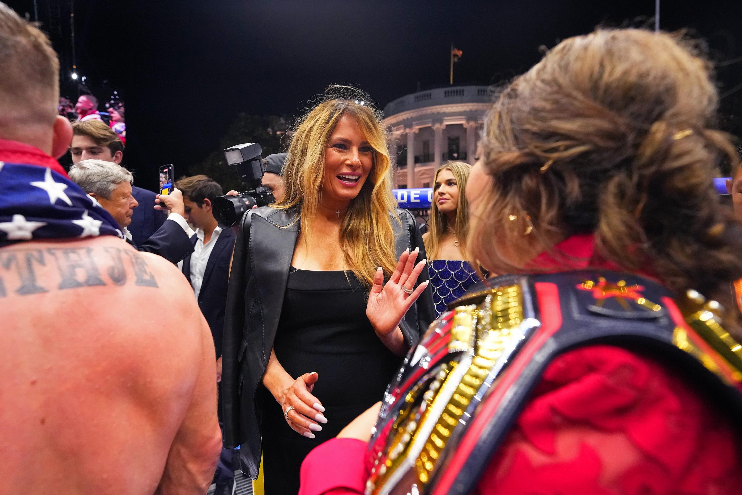 Surrounded by guests near the ring, Melania Trump gestures during a conversation. Her long highlighted hair and defined facial features stand out against the busy backdrop.