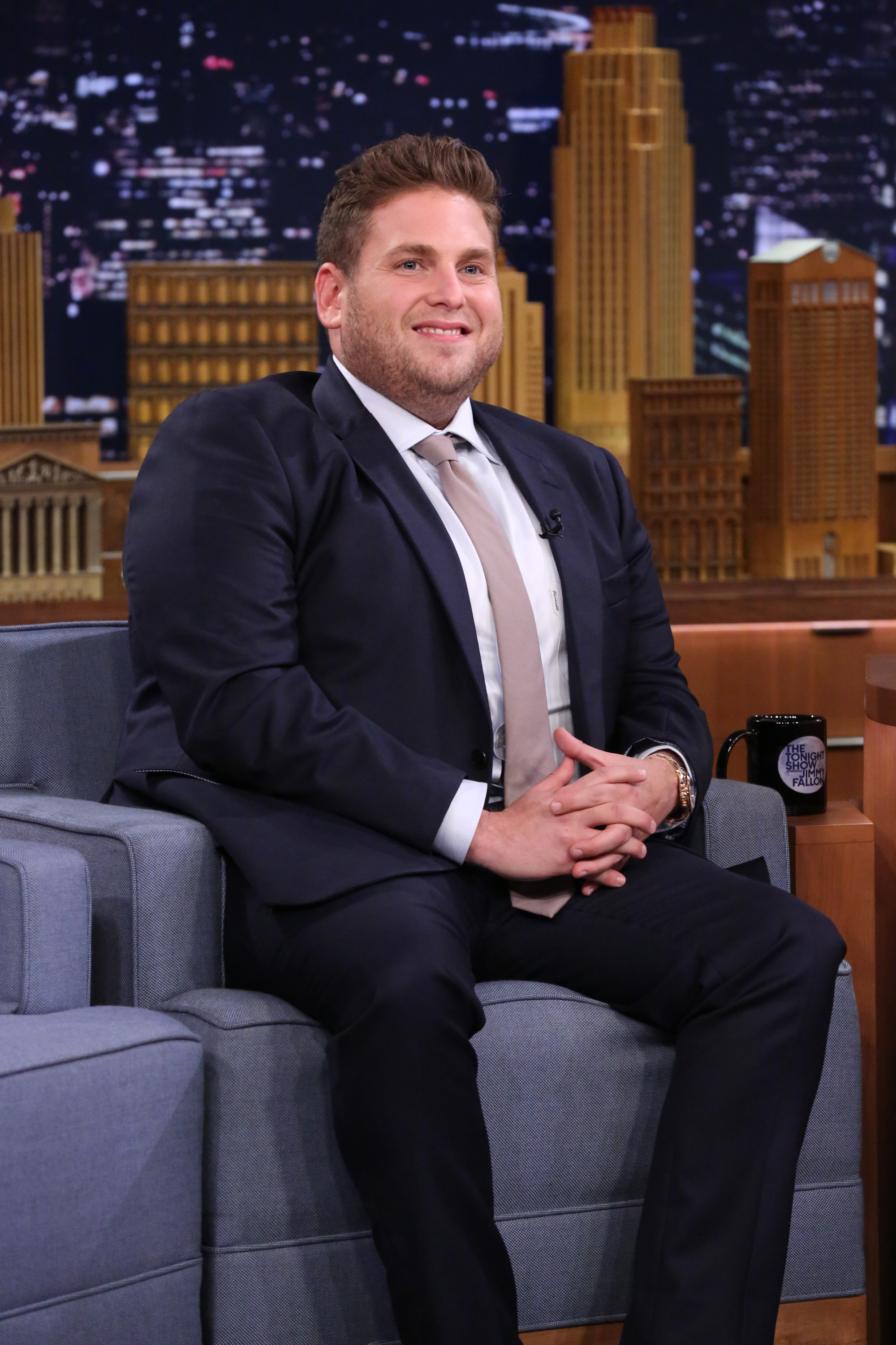 Jonah Hill appears on 'The Tonight Show Starring Jimmy Fallon' in a dark suit and beige tie, seated comfortably with his hands clasped. Smiling during the interview, he wears a gold watch and sits beside a signature show mug, with the New York City skyline set lit behind him.