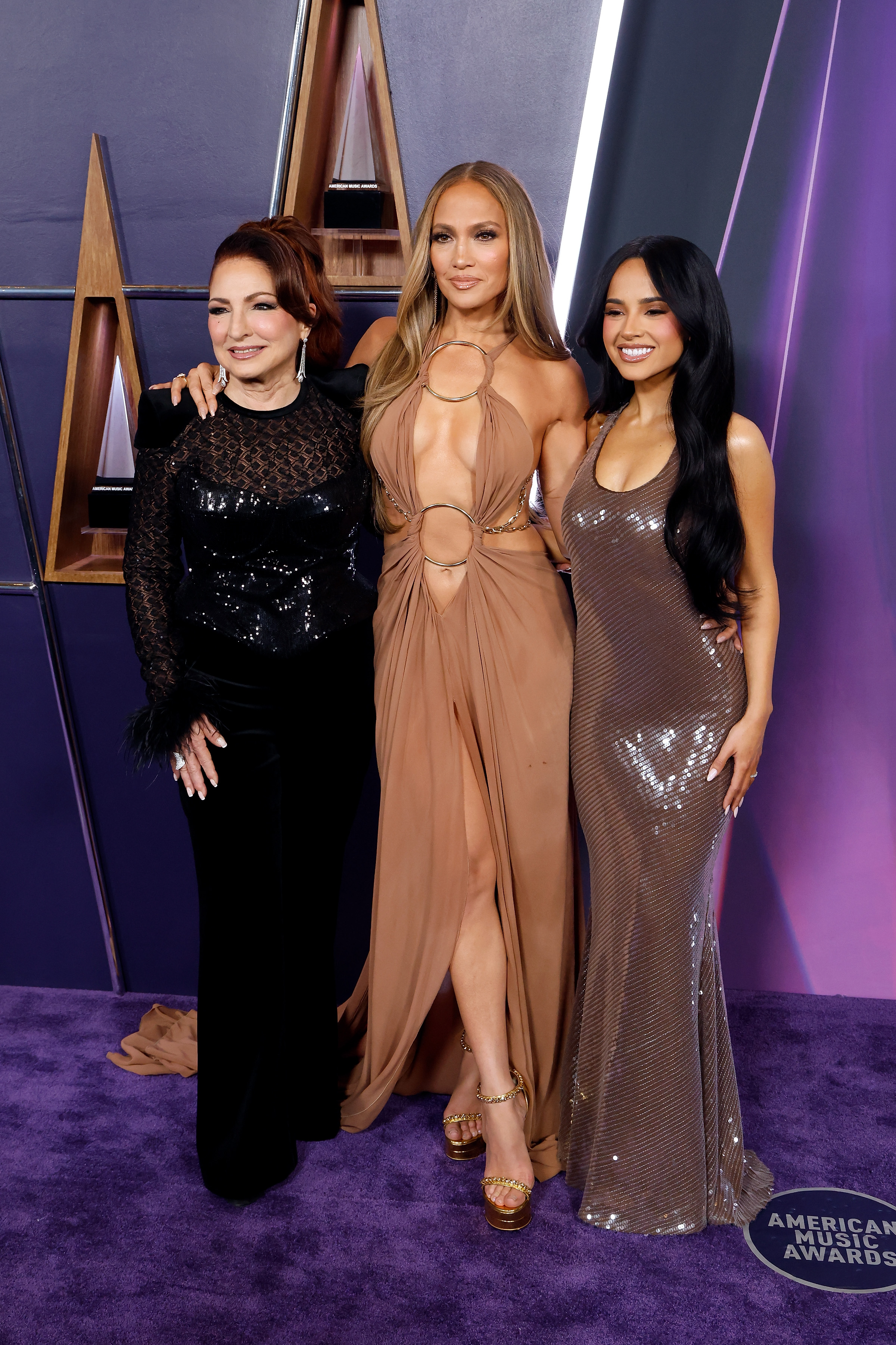 Gloria Estefan, Jennifer Lopez, and Becky G smile together during the 2025 American Music Awards, with Lopez standing in the center in her daring DEFAÏENCE gown.