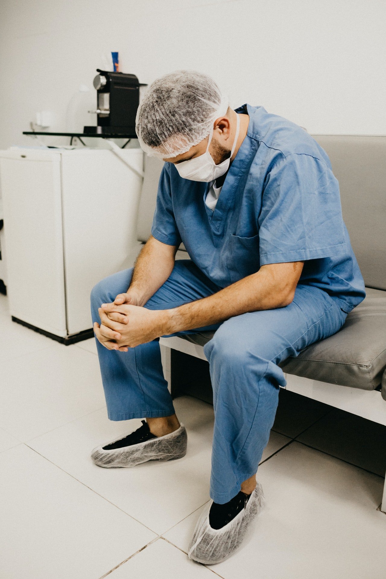 Médico con su vestimenta para cirugía sentado en un sofá de hospital mientras mira el suelo. | Foto: Pexels