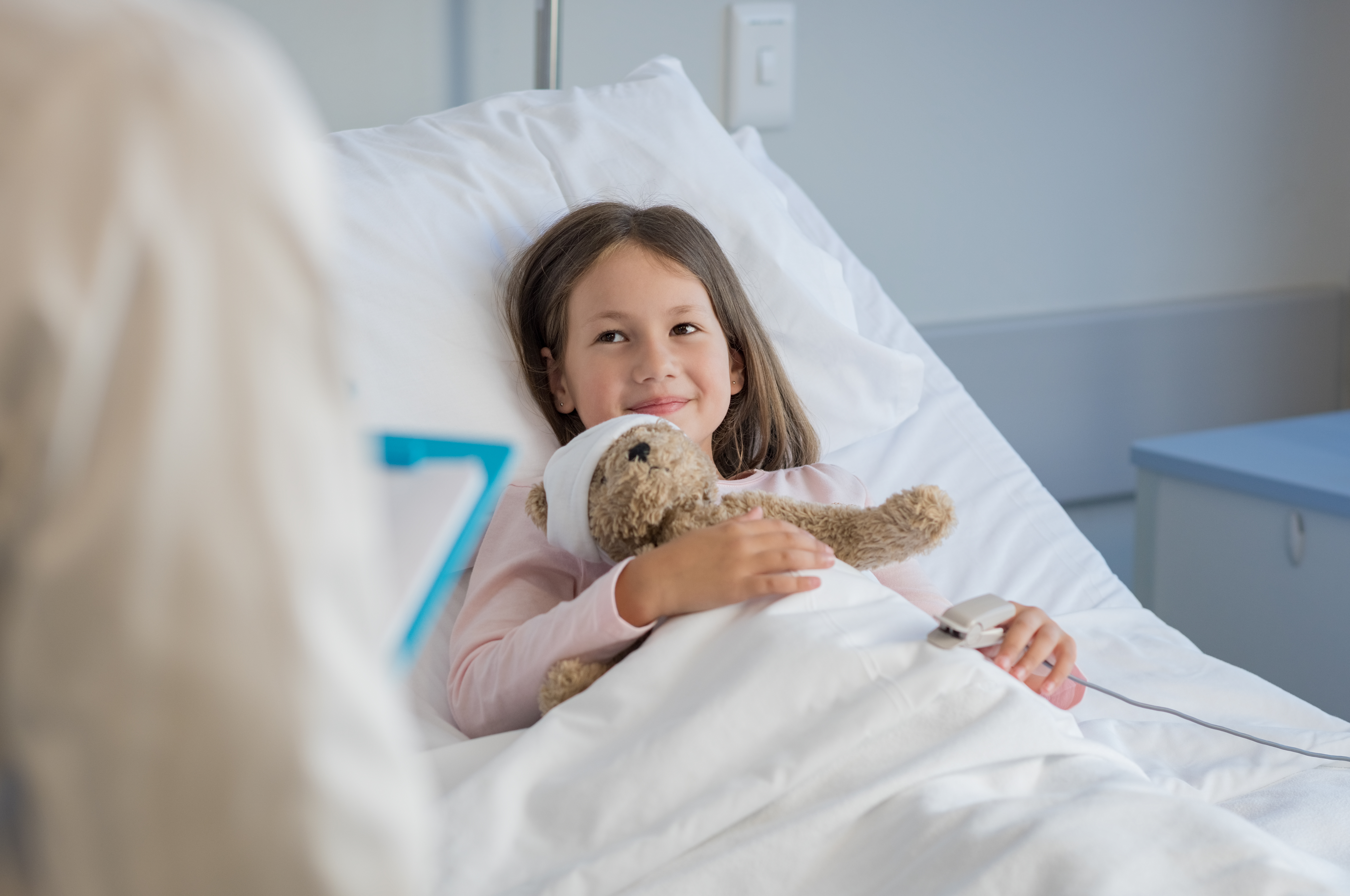Smiling little girl with oxygen saturated probe resting on hospital bed. Girl patient looking at doctor with a smile. Child and doctor at medical clinic | Source: Shutterstock