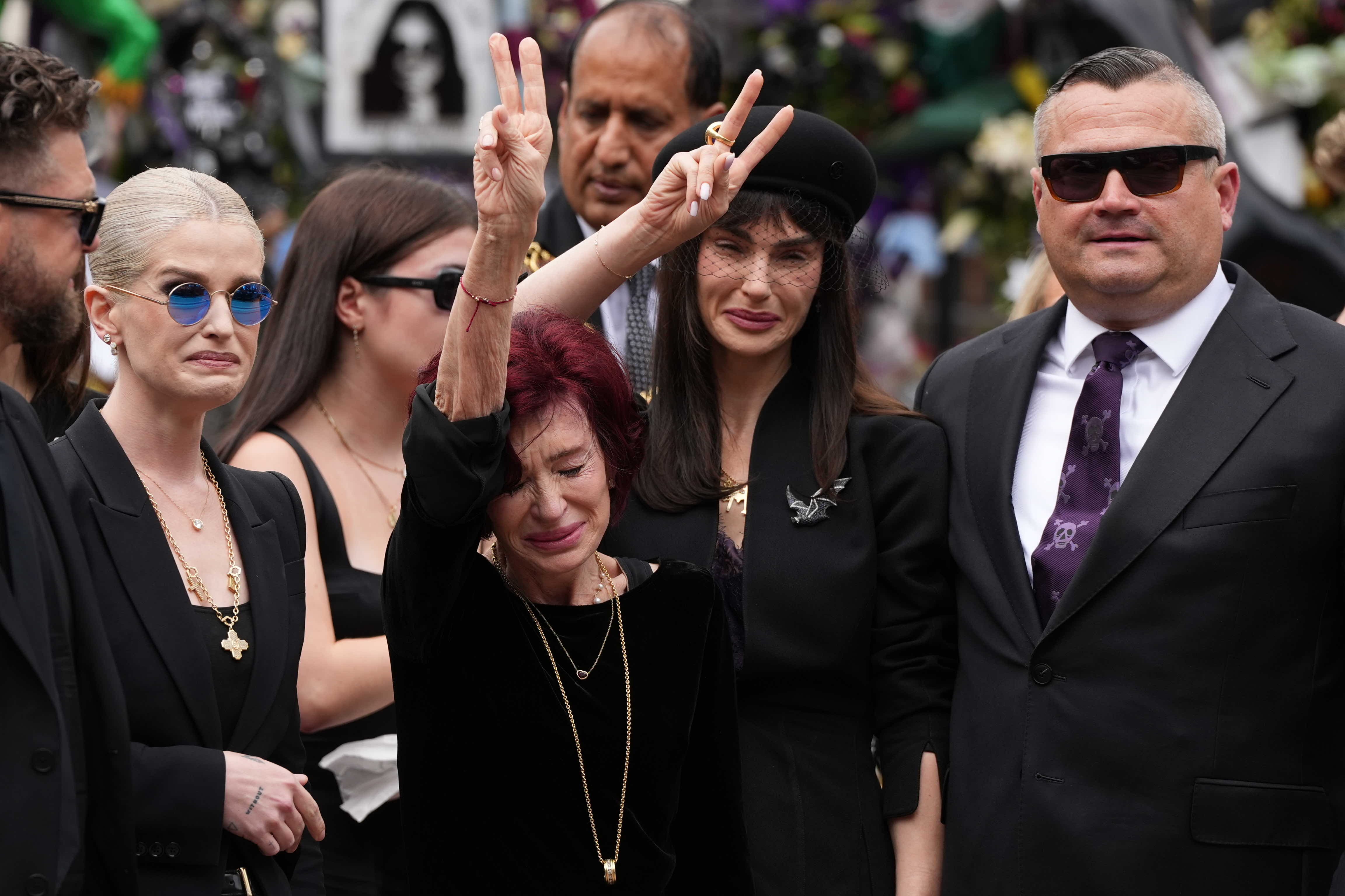 Her eyes shut tightly in grief and surrounded by loved ones at Ozzy Osbourne's funeral, Sharon Osbourne raises her arms in a peace sign. Ozzy and Sharon's daughter, Aimee also does the peace sign