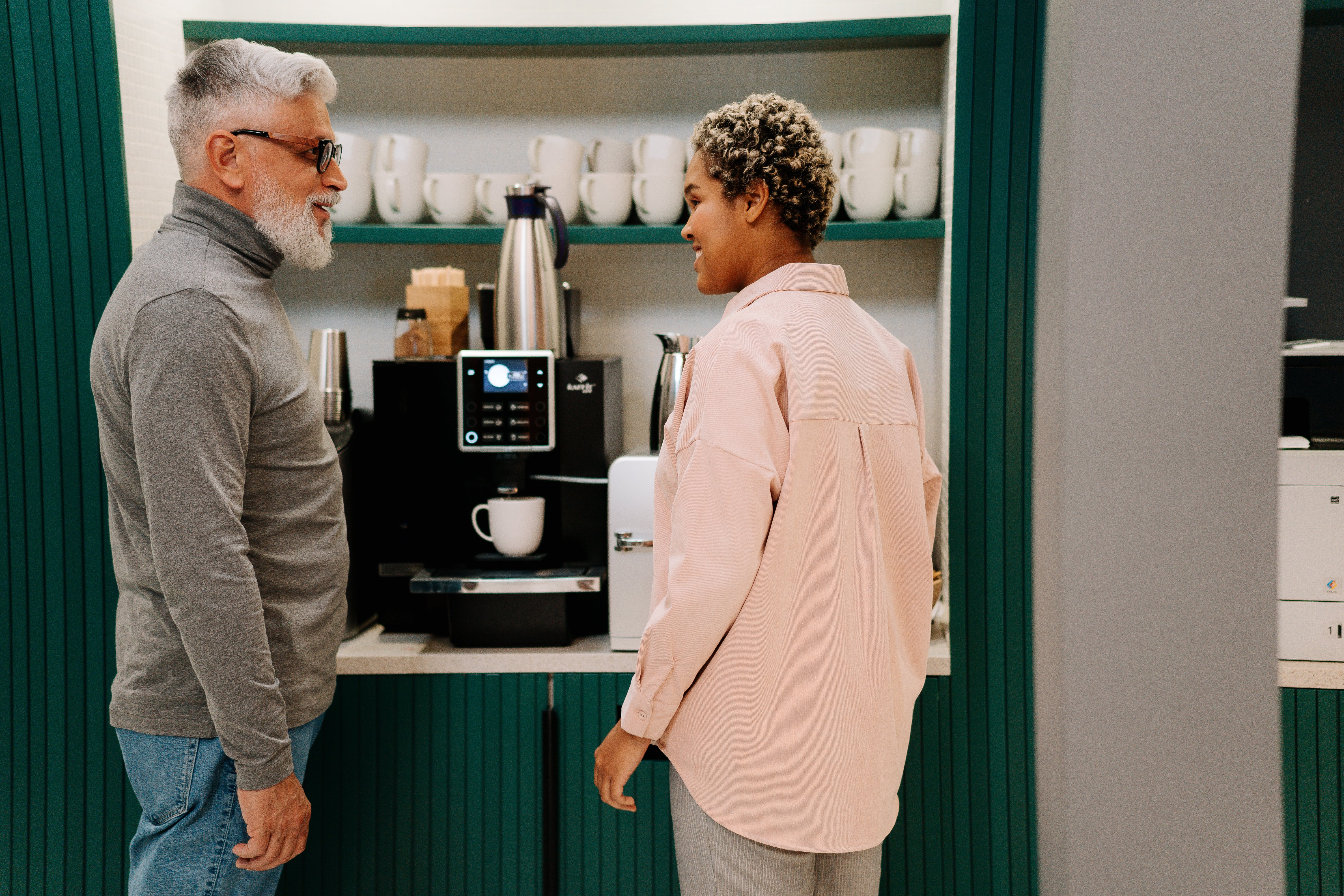 An older man talking to a younger woman while standing in front of a coffee machine | Source: Pexels